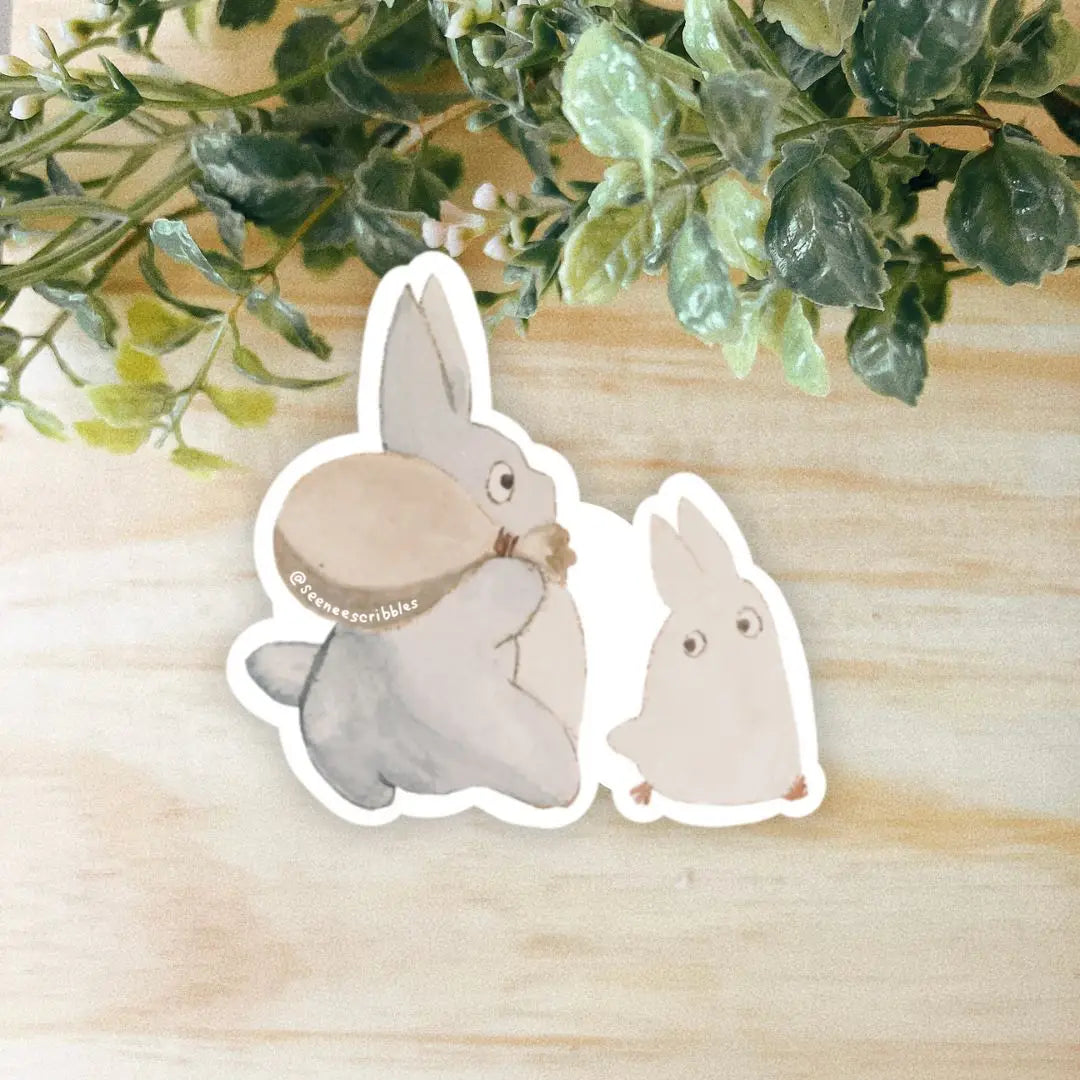 Two illustrated rabbits, one gray and the other white, sit together on a light wooden surface with green foliage above. The Seenee Scribbles Studio Ghibli Inspired Mini Totoros Stickers bring a touch of Studio Ghibli charm to your space.