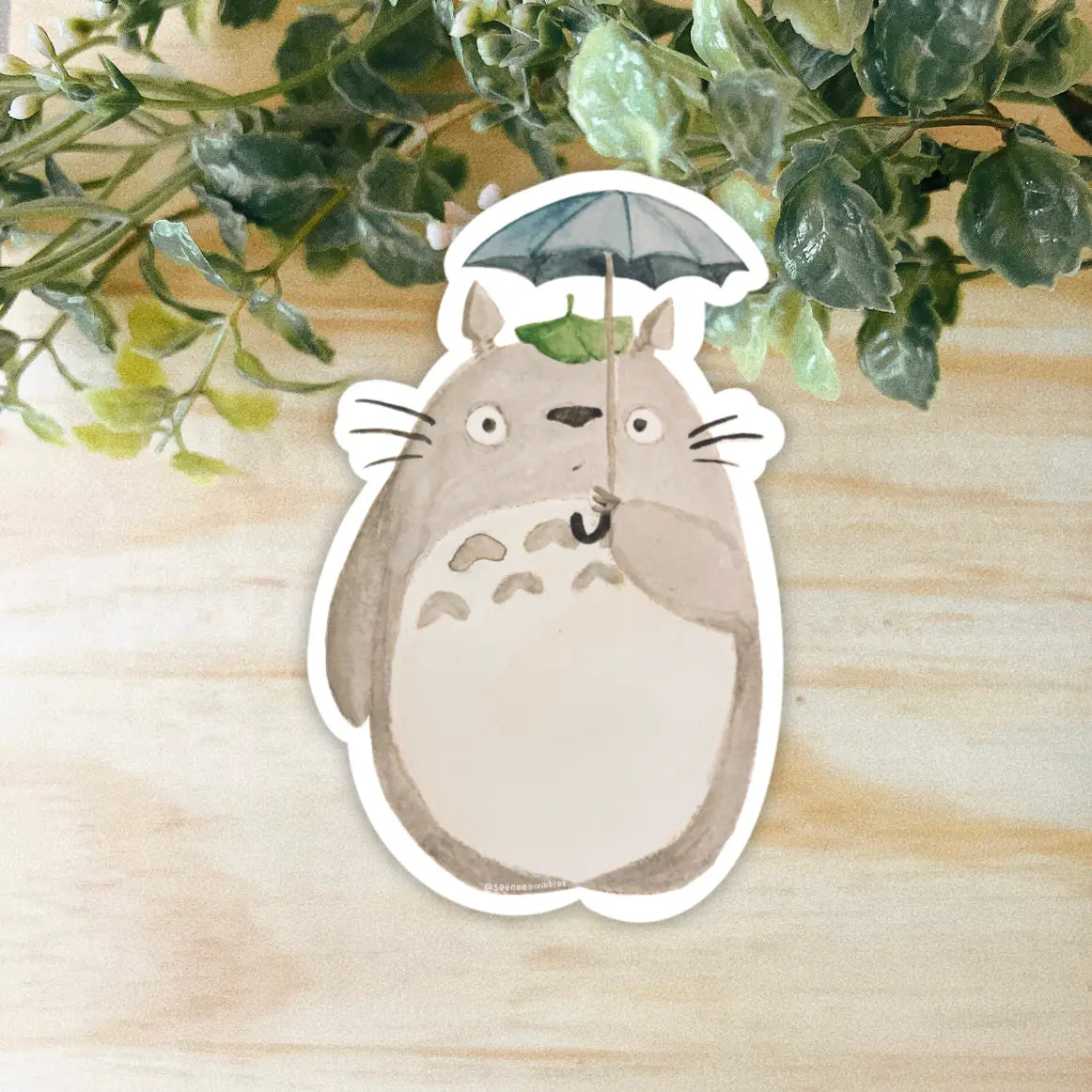 An adorable Totoro vinyl sticker, featuring a cartoon-like, chubby grey creature with a white belly holding a tiny umbrella and surrounded by green leaves, designed for fans of Studio Ghibli. Get your Seenee Scribbles Studio Ghibli Inspired Totoro Sticker today!