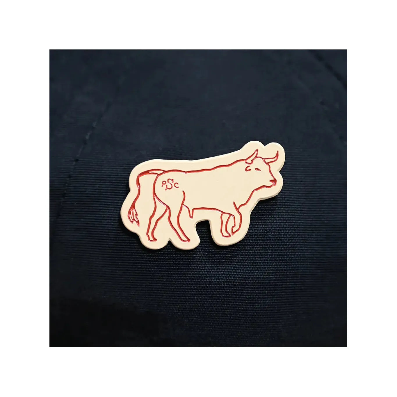 Illustration of a red-outlined cow on a cream-colored background sticker, showing the letters "PSC" on its hindquarters. The design evokes the charm of Sendero Provisions' The Ferdinand Pin with its detailed artistry and single clutch backing.