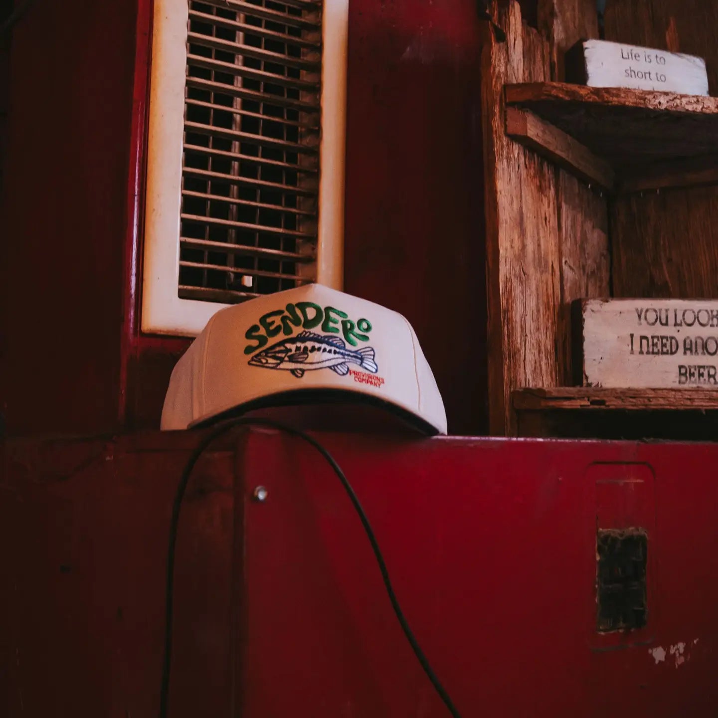 Sendero Lunker Fish Cap in white with colorful fish design on red table, displayed near store shelves and an air vent.