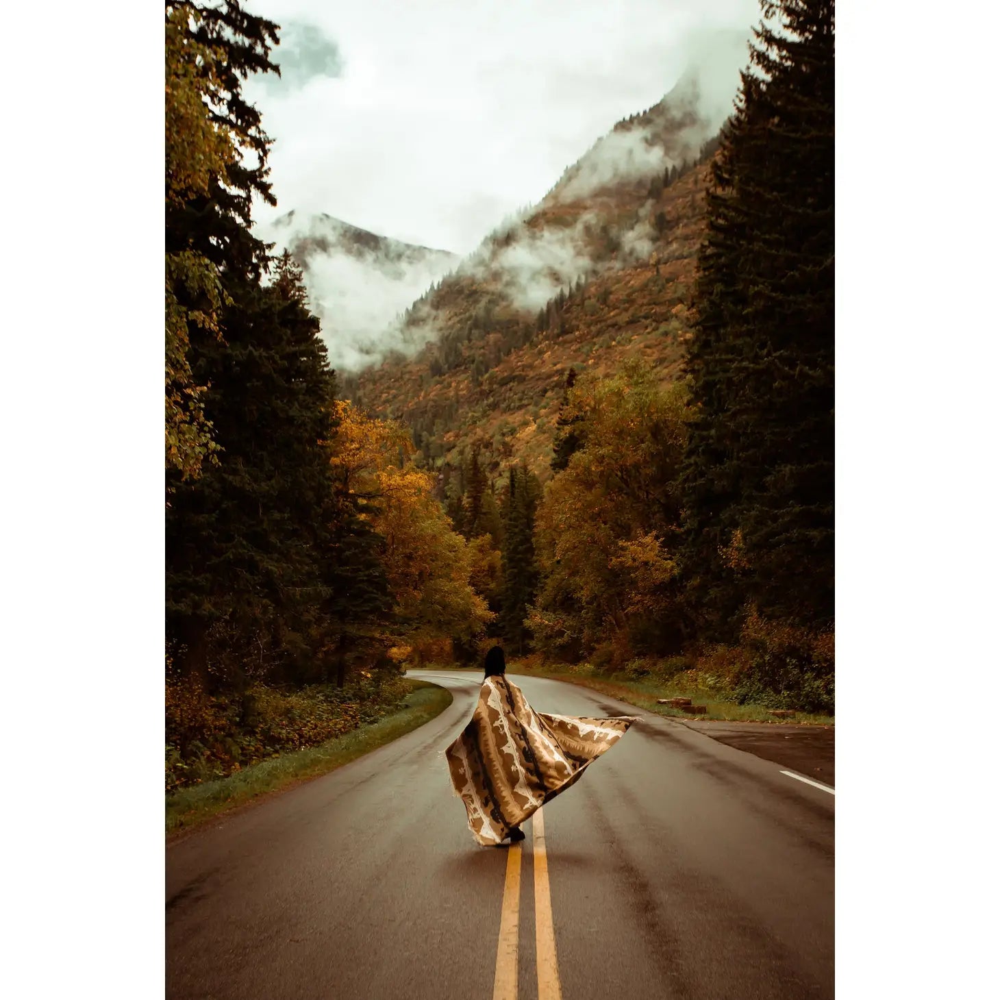 A person draped in the Sackcloth & Ashes Smokey Bear Blanket - Smokey Roam walks along a road through a lush forest, with mountains and clouds forming a picturesque backdrop.