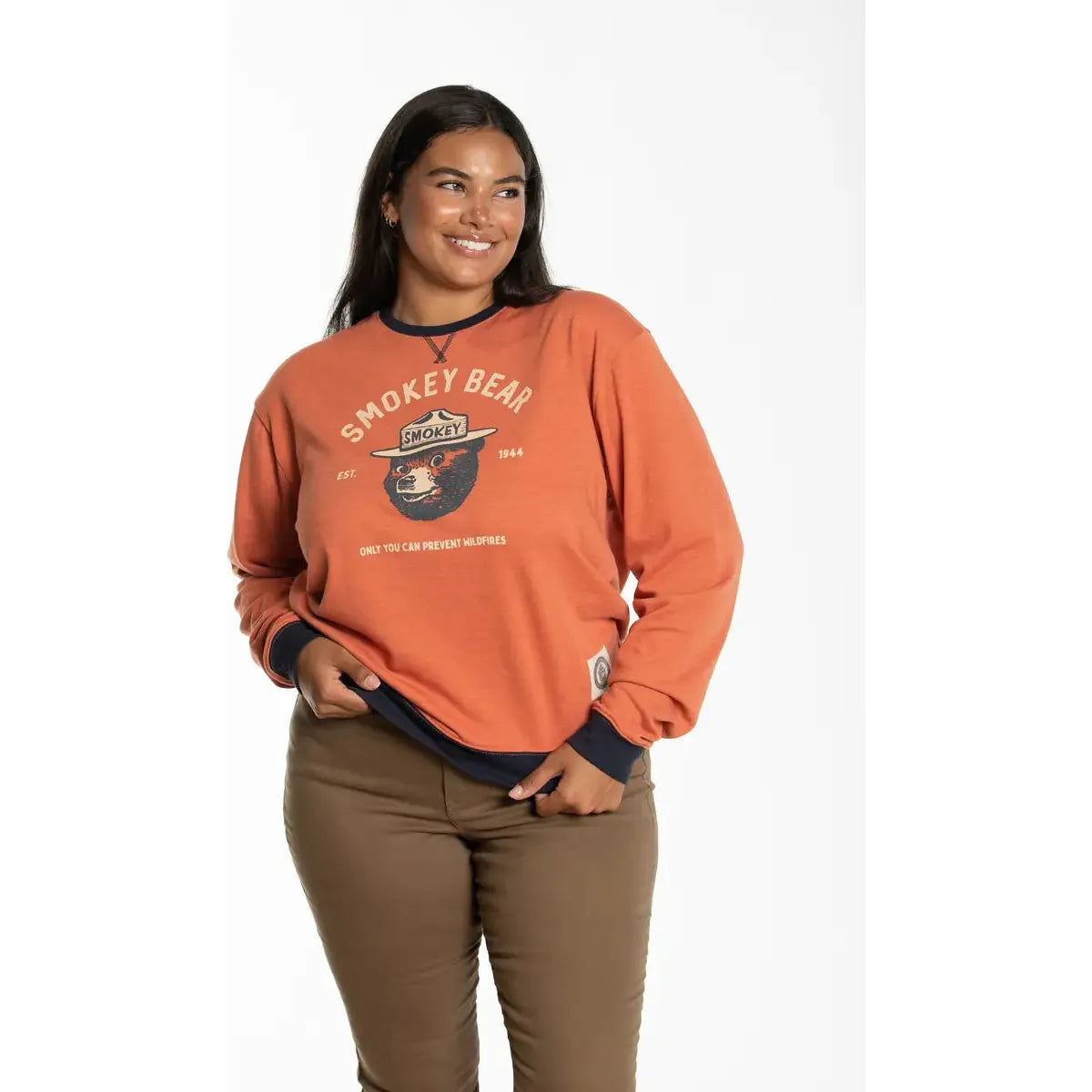 A person smiling, wearing The Landmark Project Smokey Bear Heritage Varsity Orange Sweatshirt, which features an illustration of Smokey Bear and text about preventing wildfires. Proudly made in Guatemala from BCI Cotton, ensuring a sustainable choice.