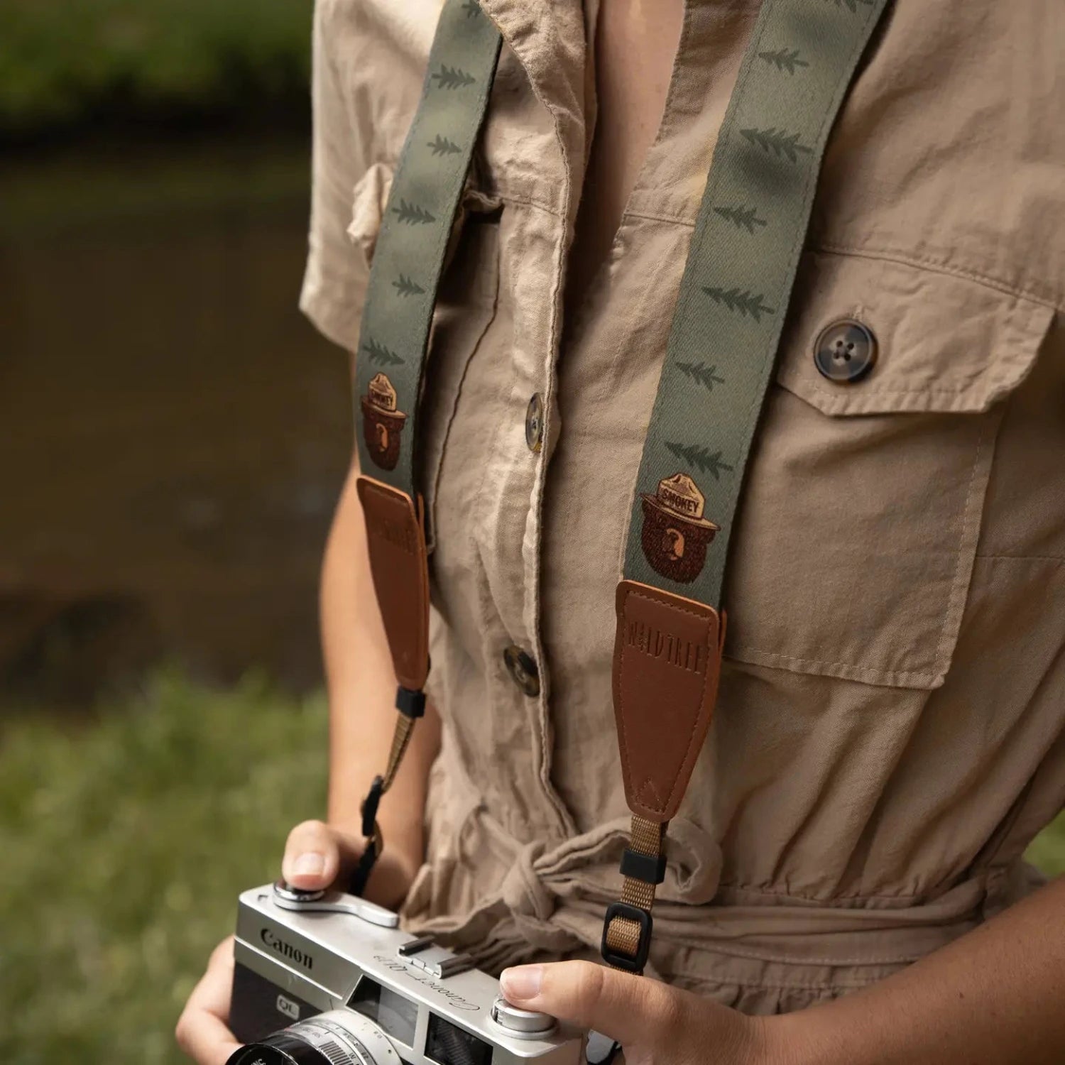 Person in tan jumpsuit holds camera with Wildtree Smokey Bear strap by grassy water, outdoor photography gear scene.