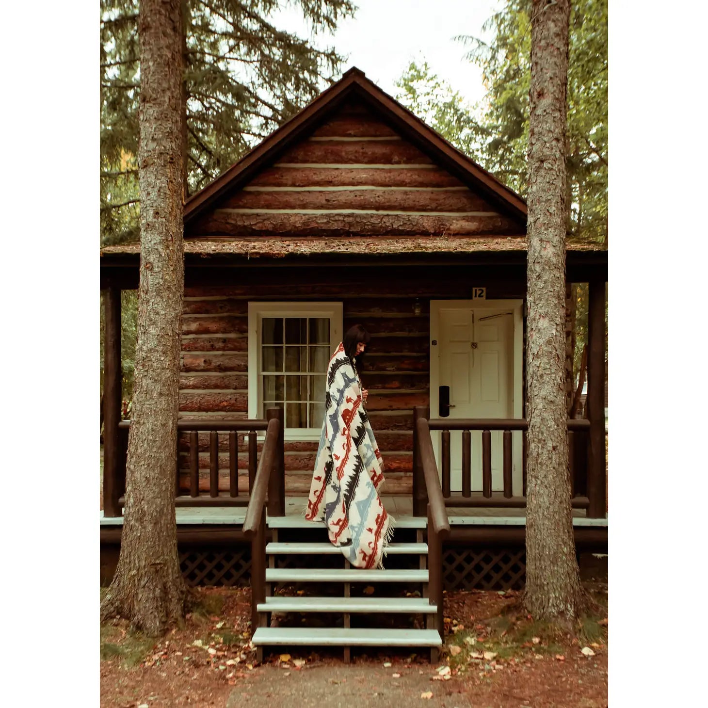 Person wrapped in a Sackcloth & Ashes Smokey Bear Blanket - Smokey Ramble stands on the steps of a rustic log cabin surrounded by trees, embodying a reminder for wildfire prevention.