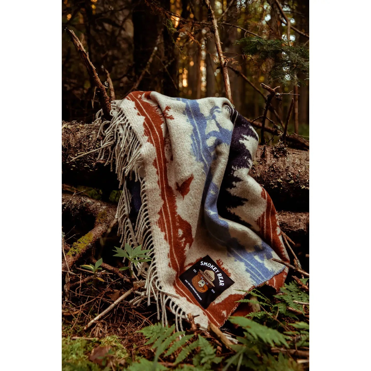 A cozy Sackcloth & Ashes Smokey Bear Blanket - Smokey Ramble with a colorful nature pattern draped over fallen logs in a lush forest, promoting wildfire prevention.