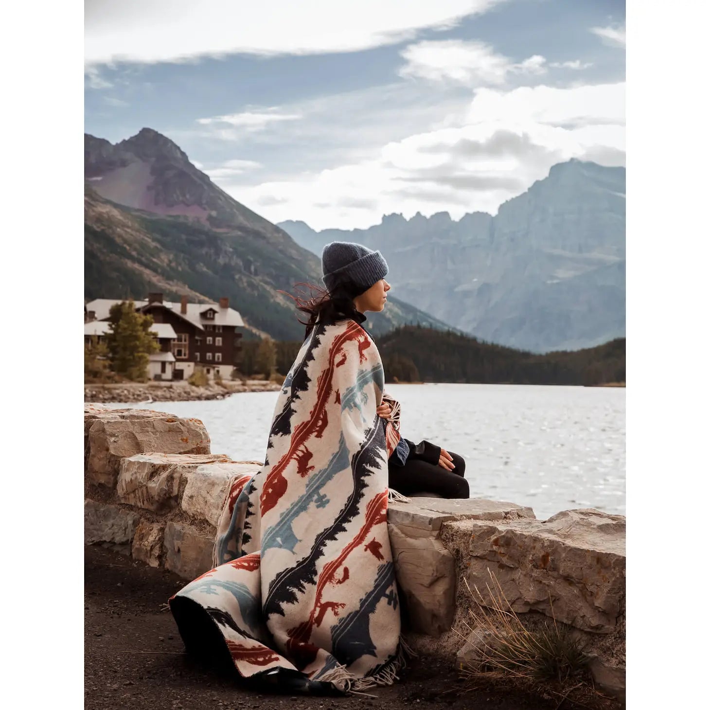 Person in a beanie sitting on a stone wall by a lake, wrapped in a Sackcloth & Ashes Smokey Bear Blanket - Smokey Ramble, with mountains and a lodge in the background.