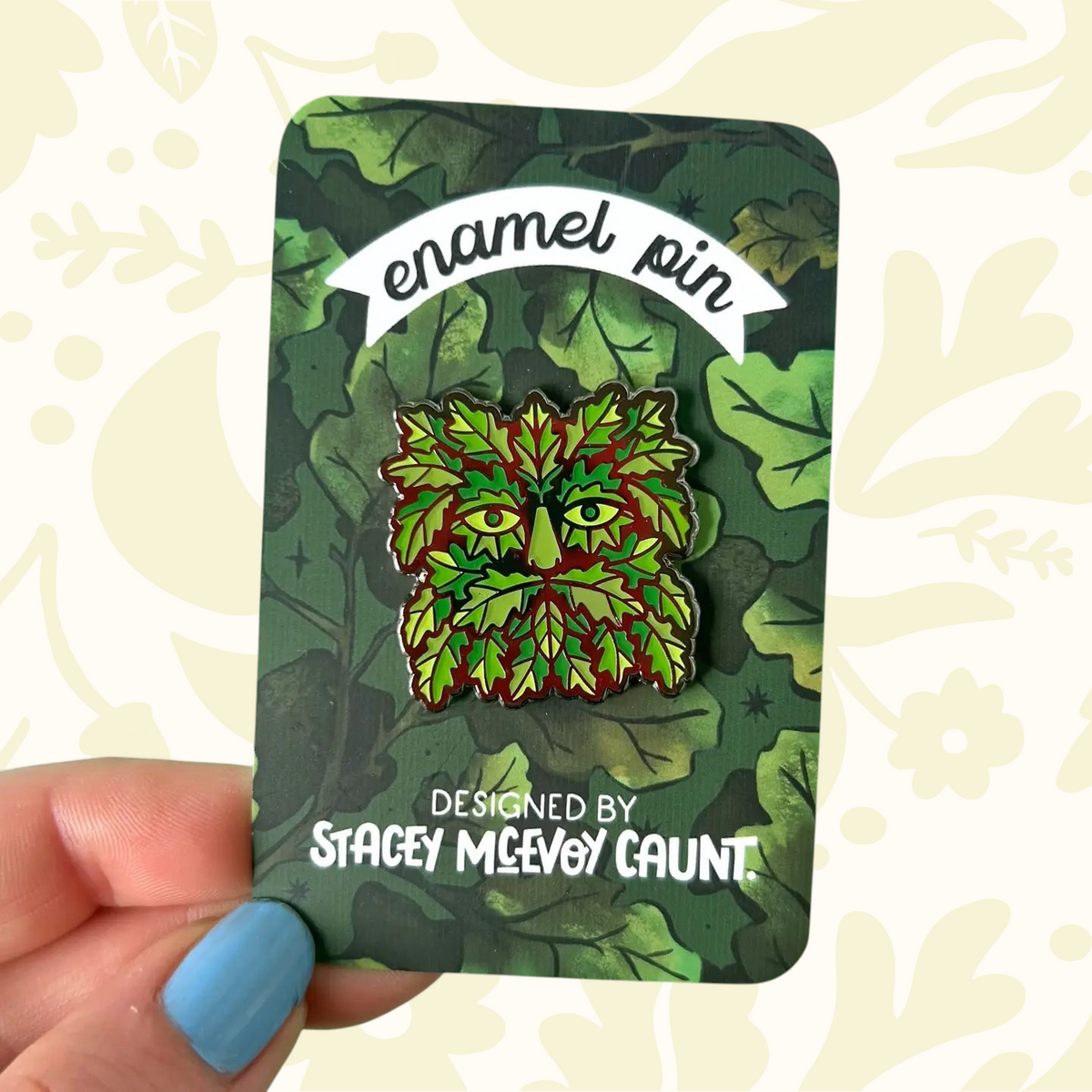 Hand displays Stacey McEvoy Caunt Green Man enamel pin on leafy card, perfect for nature, plant, and folklore lovers.