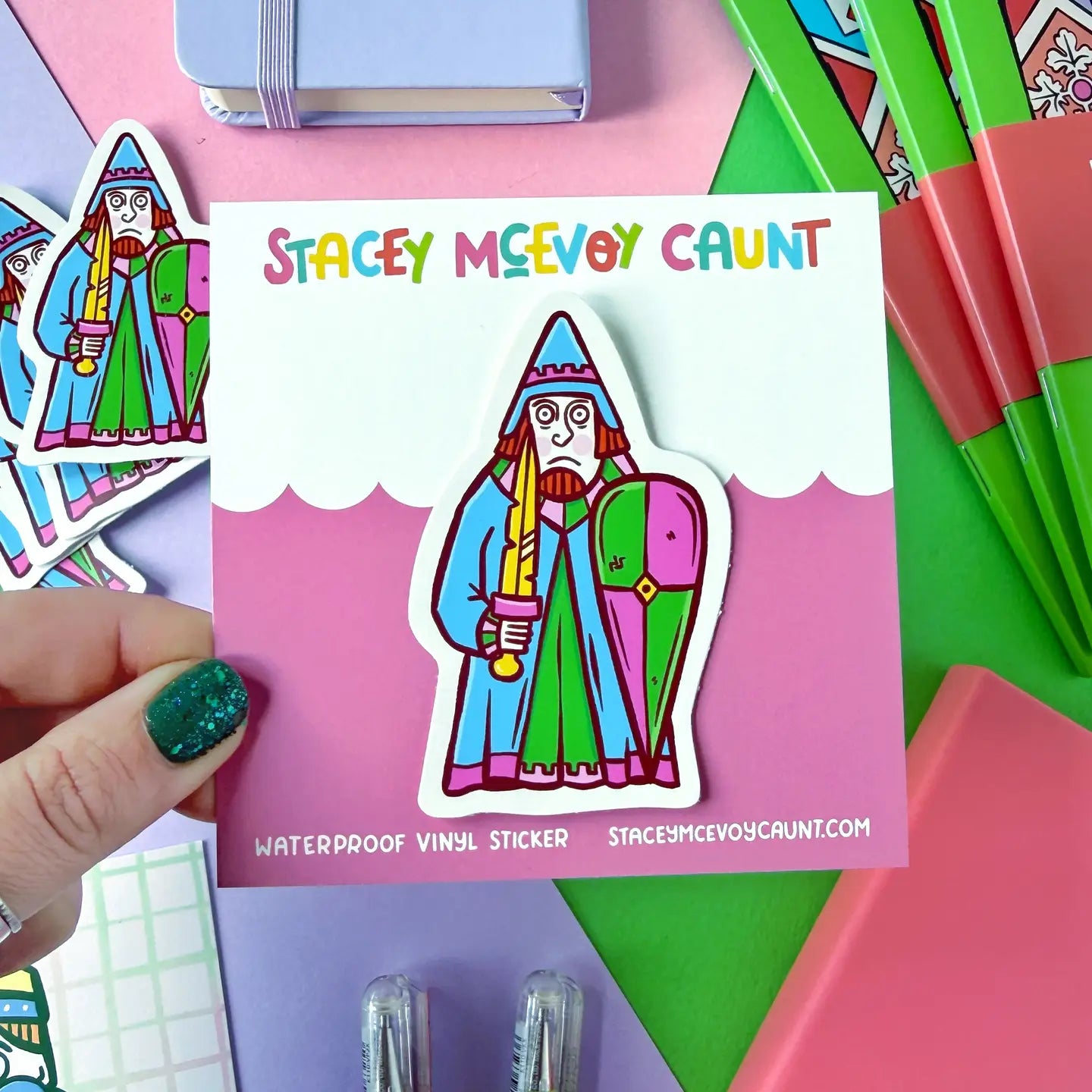 Hand displays Stacey McEvoy Caunt medieval rook Lewis chessmen vinyl sticker, branded card, plus colorful stationery in background.