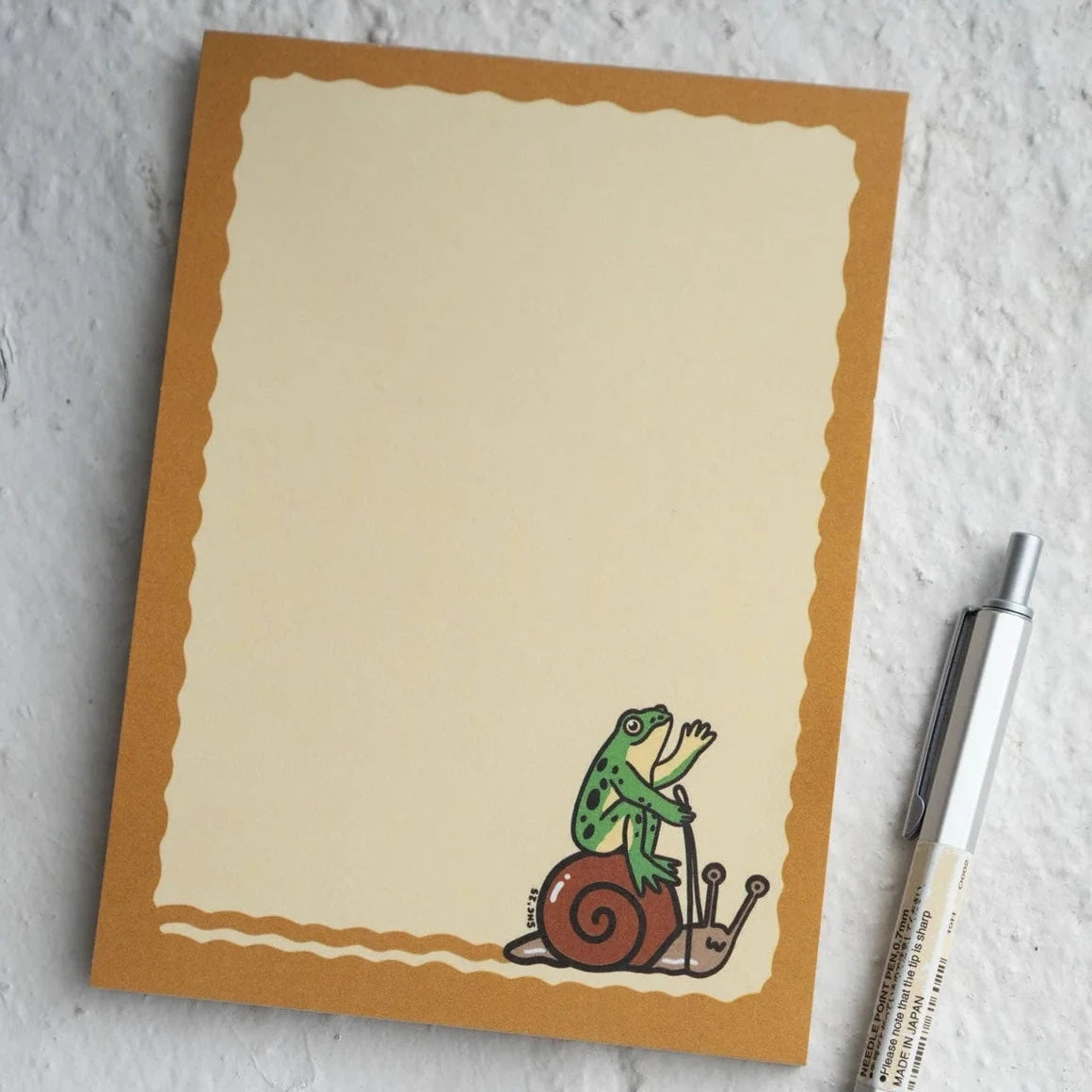 Stay Home Club Frog & Snail Ride Notepad featuring a cute frog riding a snail, brown border, and nature-inspired stationery design.