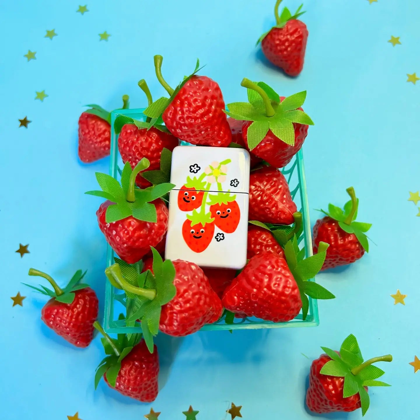 Small basket of artificial strawberries and The Peach Fuzz Sweet Strawberry Lighter on blue background with gold stars.