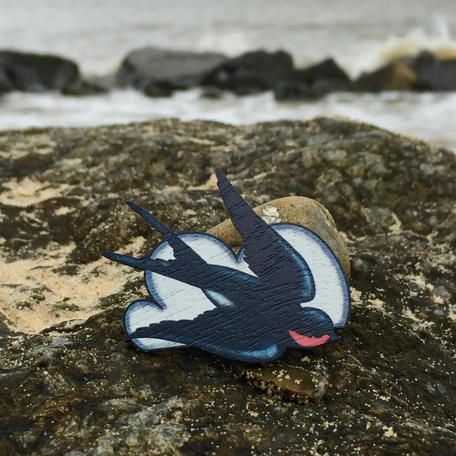 The Su Owen Swallows Brooch features a wooden pin in the shape of a flying swallow, with black and white wings and a red neck, crafted from sustainable walnut by Su Owen, set against a bright blue background.