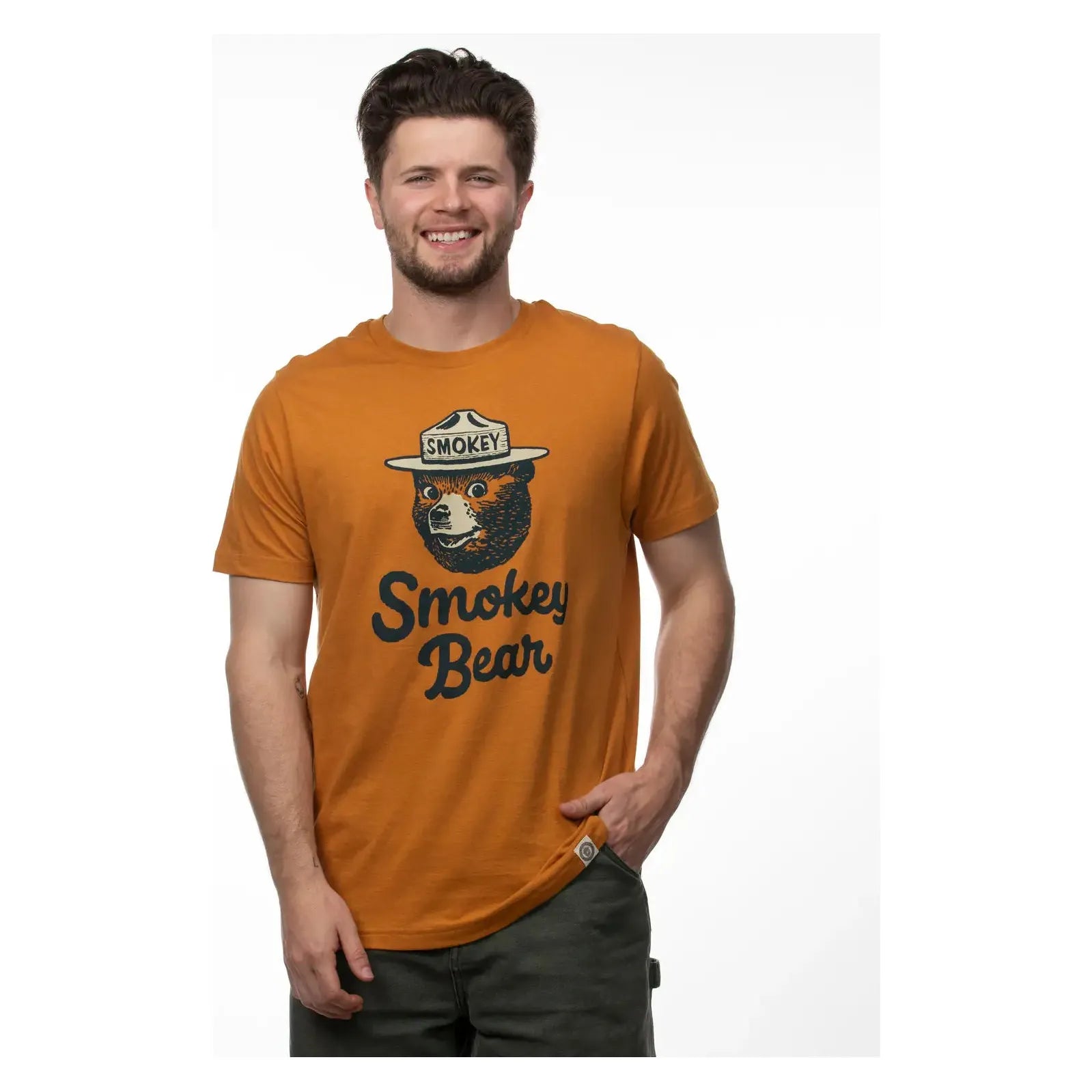 A bearded man wearing The Landmark Project Heritage Smokey Bear T-Shirt in Sundial, featuring vintage colors and the text "Smokey Bear," smiles at the camera.