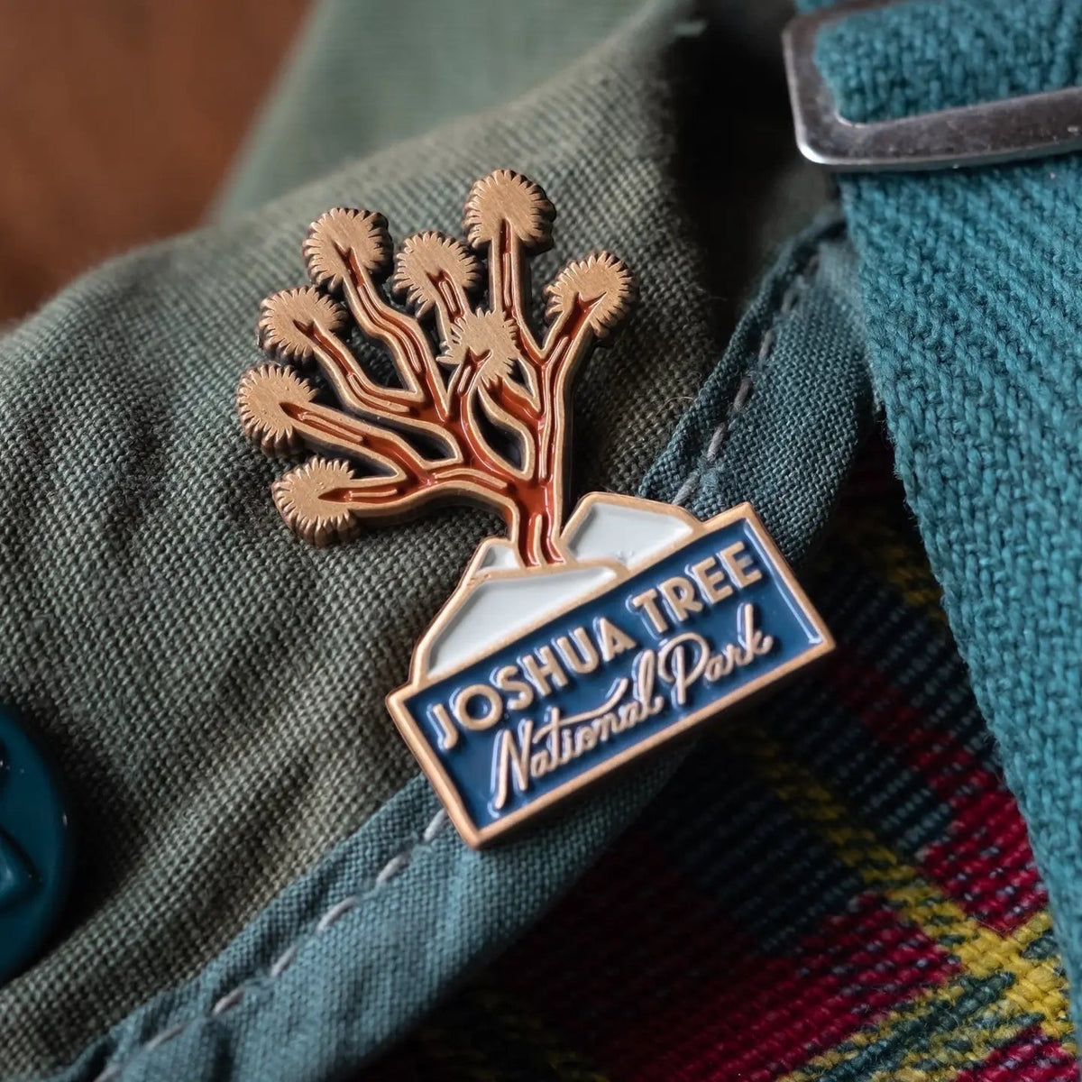 The Landmark Project Joshua Tree enamel pin on green fabric with a blue button and plaid, perfect for nature lovers and collectors.