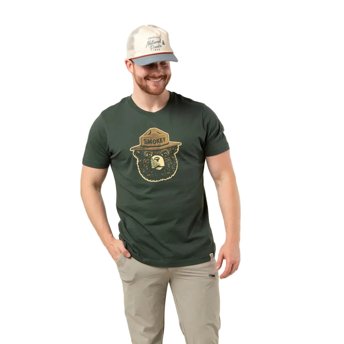 Smiling man in Smokey Bear Logo Pine T-Shirt by The Landmark Project, light pants and cap, standing on a white background.