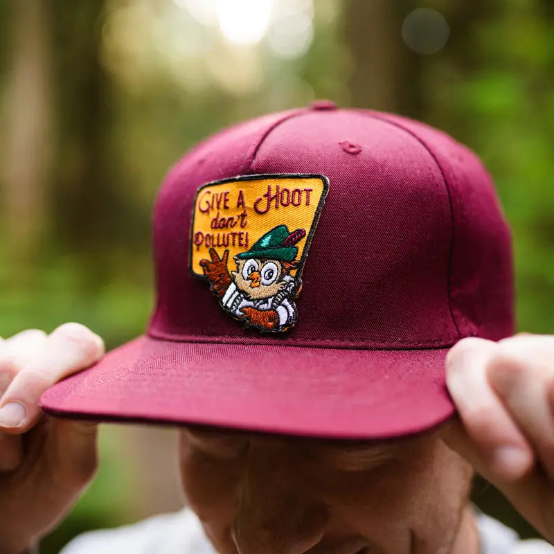 Person fixes The Landmark Project Woodsy Owl Give A Hoot Don’t Pollute patch on maroon hat—eco-friendly cap accessory.