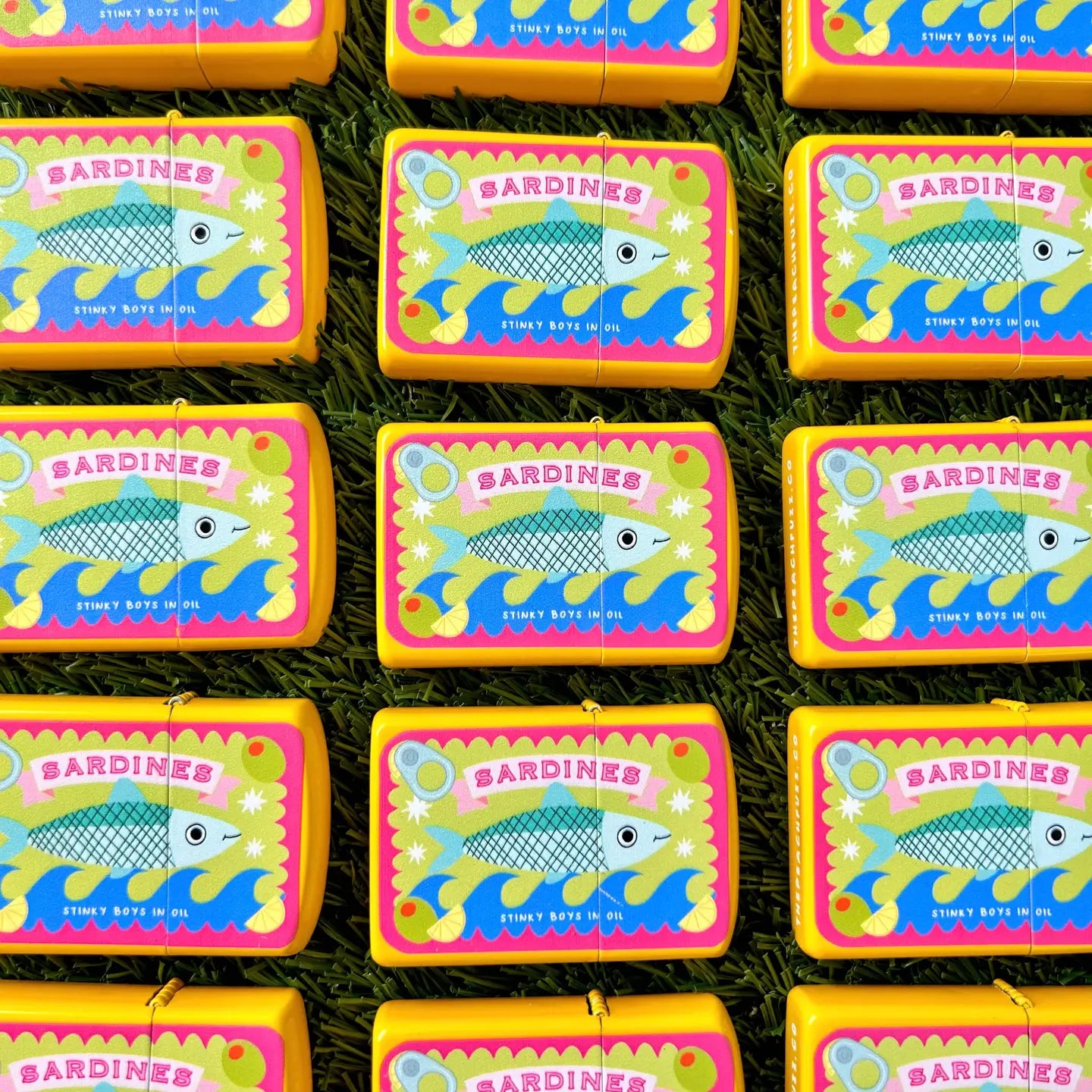 The Peach Fuzz Tinned Sardine Refillable Lighters with fish designs are lined up on green grass, unique and eco-friendly.