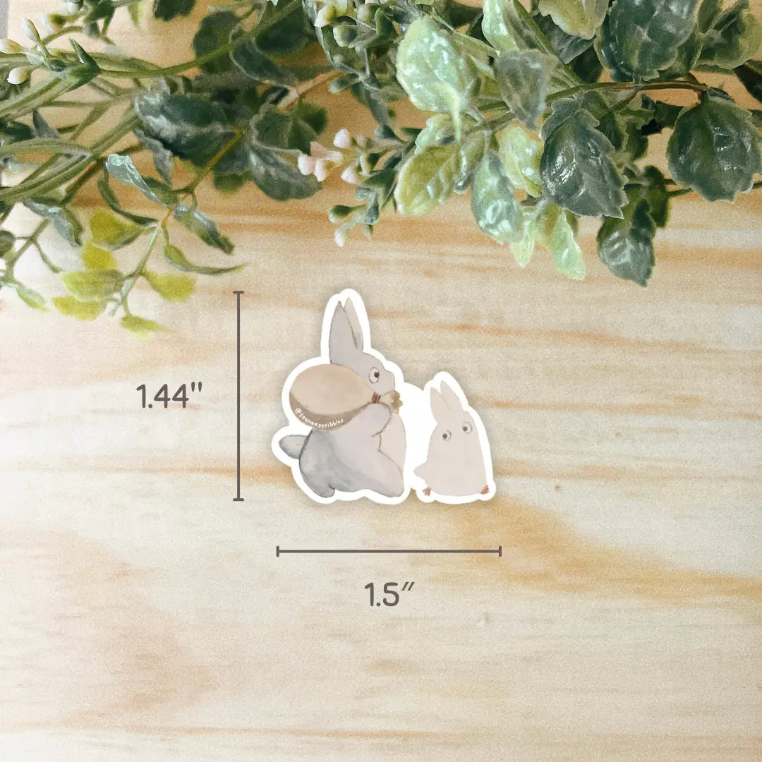 Two illustrated rabbits, one gray and the other white, sit together on a light wooden surface with green foliage above. The Seenee Scribbles Studio Ghibli Inspired Mini Totoros Stickers bring a touch of Studio Ghibli charm to your space.