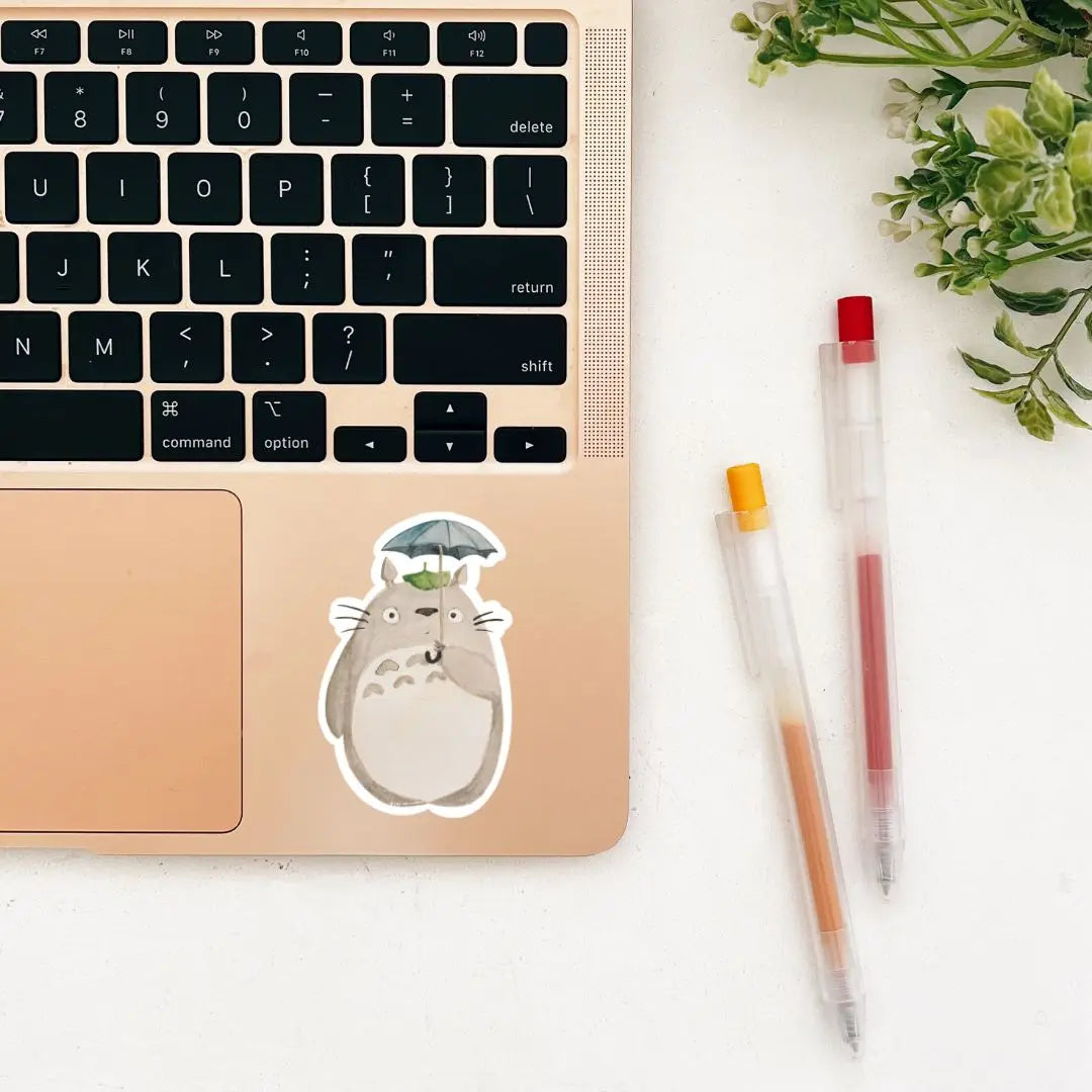 An adorable Totoro vinyl sticker, featuring a cartoon-like, chubby grey creature with a white belly holding a tiny umbrella and surrounded by green leaves, designed for fans of Studio Ghibli. Get your Seenee Scribbles Studio Ghibli Inspired Totoro Sticker today!