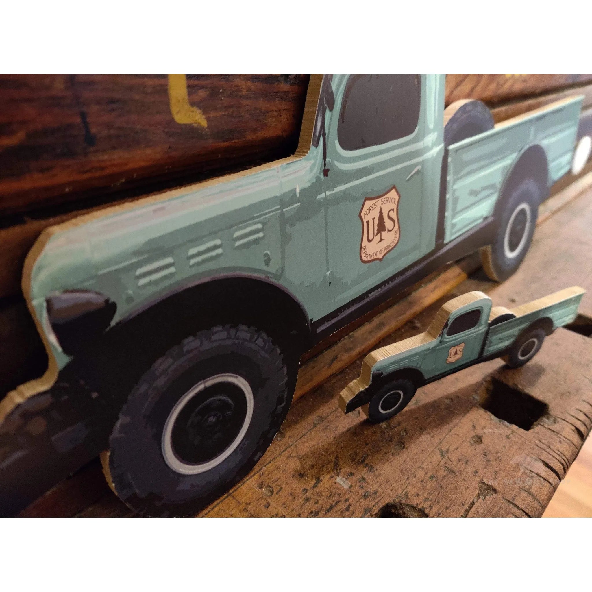 Close-up of two The Sawmill Shop ‘Made In USA’ Forest Service Trucks, crafted from wood and painted green with "U.S." logos, placed on a wooden surface. The larger truck is in the background.