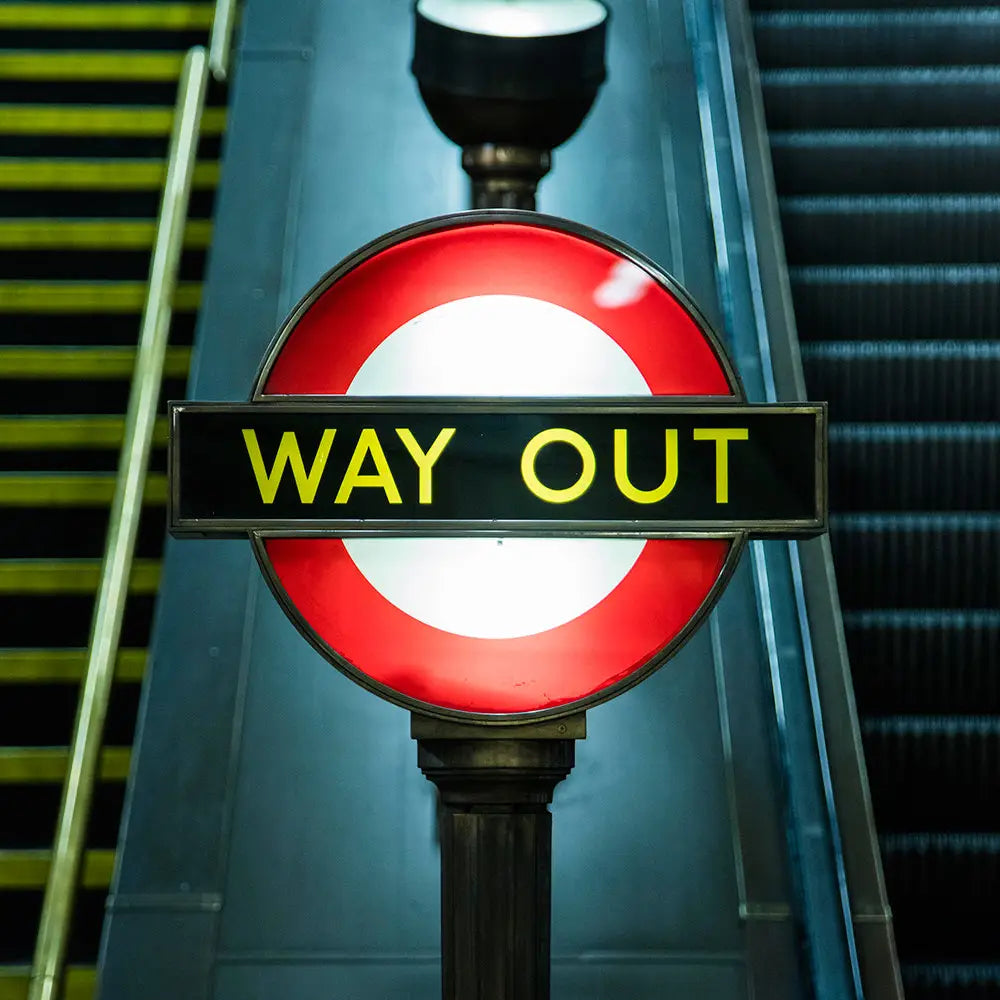 In the London Underground station, a sign displaying "WAY OUT" text at the center of a red and white circle is positioned between a staircase and an escalator, as featured in the Blue Crow Media London Underground Architecture Map.