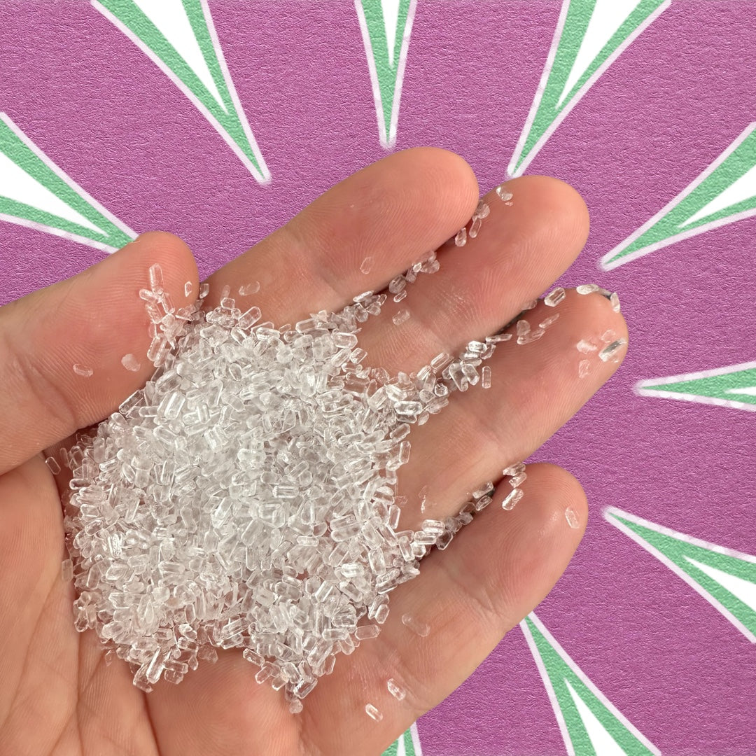 Close-up of a hand holding clear, coarse Epsom salt crystals against a bold purple and green patterned background.