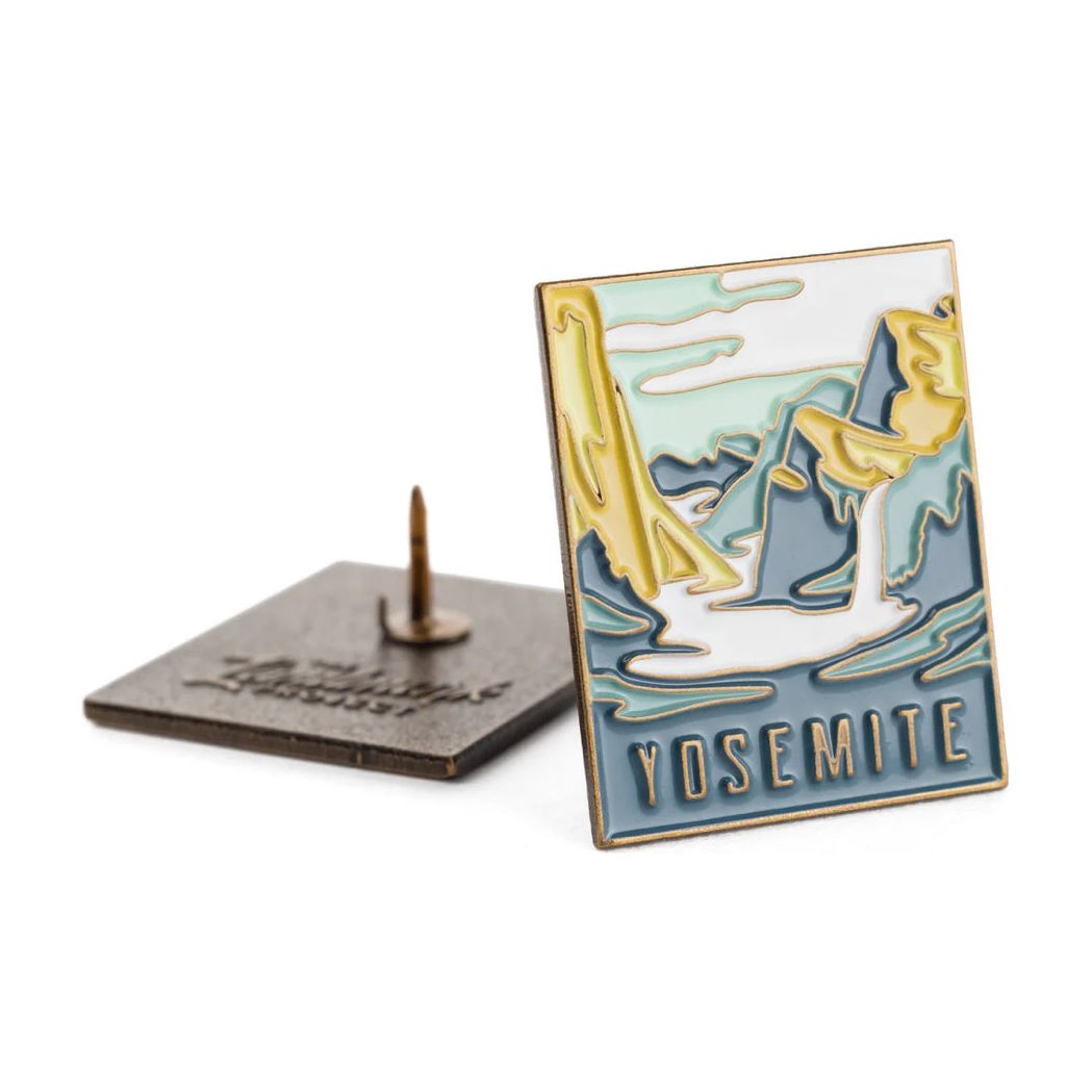 The Landmark Project Yosemite National Park Pin set includes two enamel pins showcasing a stylized depiction of Yosemite's landscape with mountains and water, alongside "Yosemite" written at the bottom. These pins are packaged in a plastic-free, sustainable manner to help protect our beautiful planet.