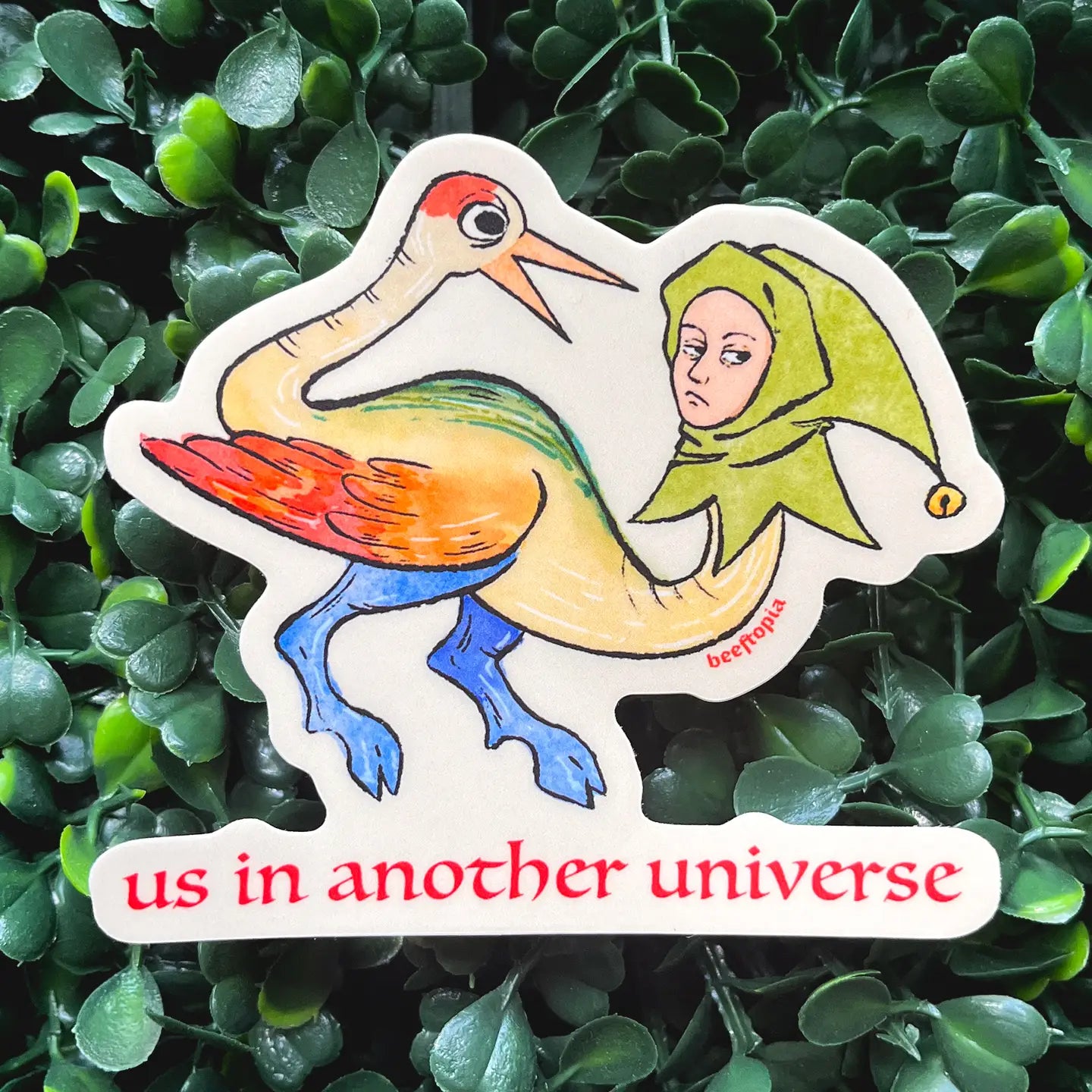 Beeftopia Us in Another Universe sticker: quirky medieval bird, human face, green hat, leaves, Beeftopia logo—funny collectible.