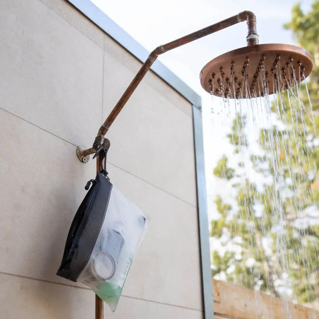 Outdoor shower with water flowing, The Pouch 3-1-1 TSA clear bag hangs on pipe, trees and gravel visible in background.