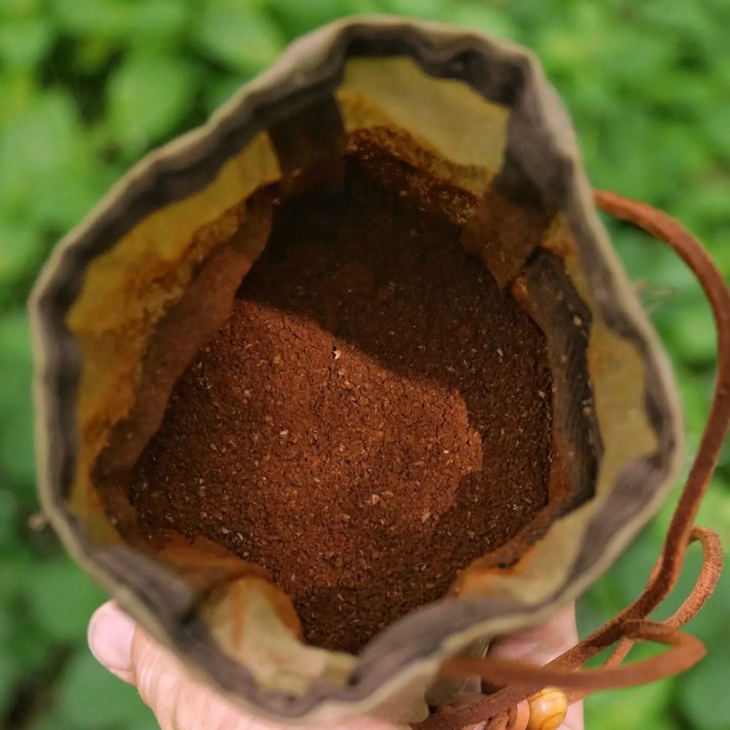Hand holding khaki green Journeyman Craft Waxed Canvas Coffee Pouch, open outdoors with leafy green background, stylish & durable.