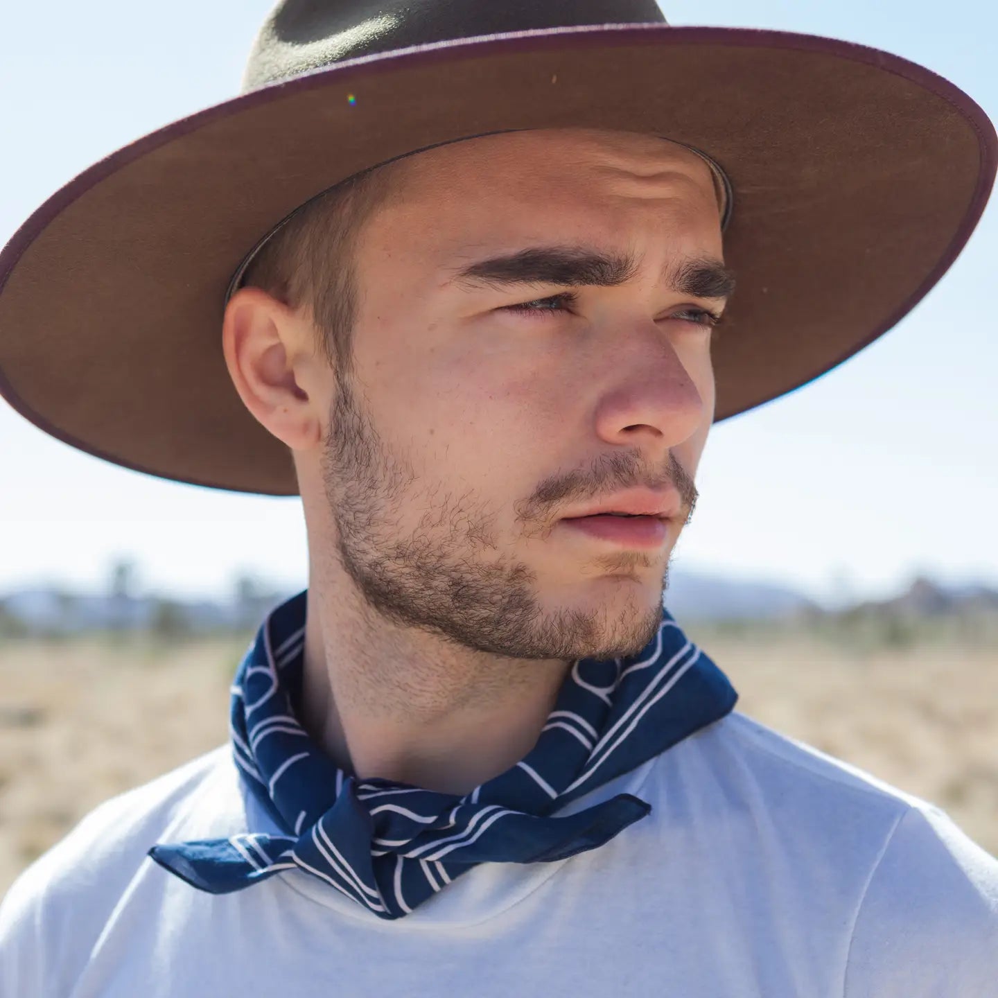 Stylish man in Bandit Bandanas “Good Luck” bandana, wide-brim hat, outdoors in sunlit, dry landscape looking sideways.