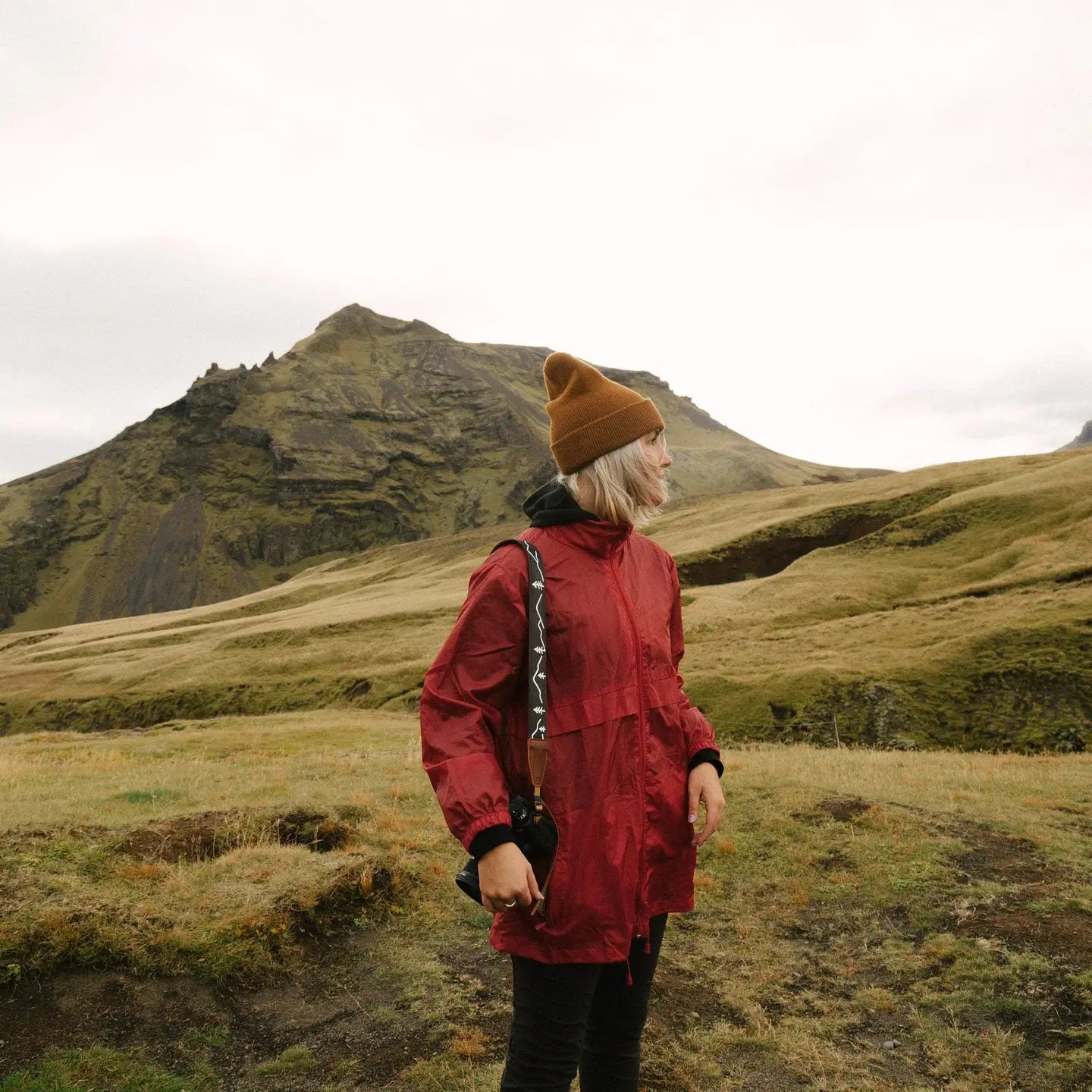 Person in red jacket with Wildtree Simple Mountain Design strap stands on grassy mountain under cloudy, overcast sky.