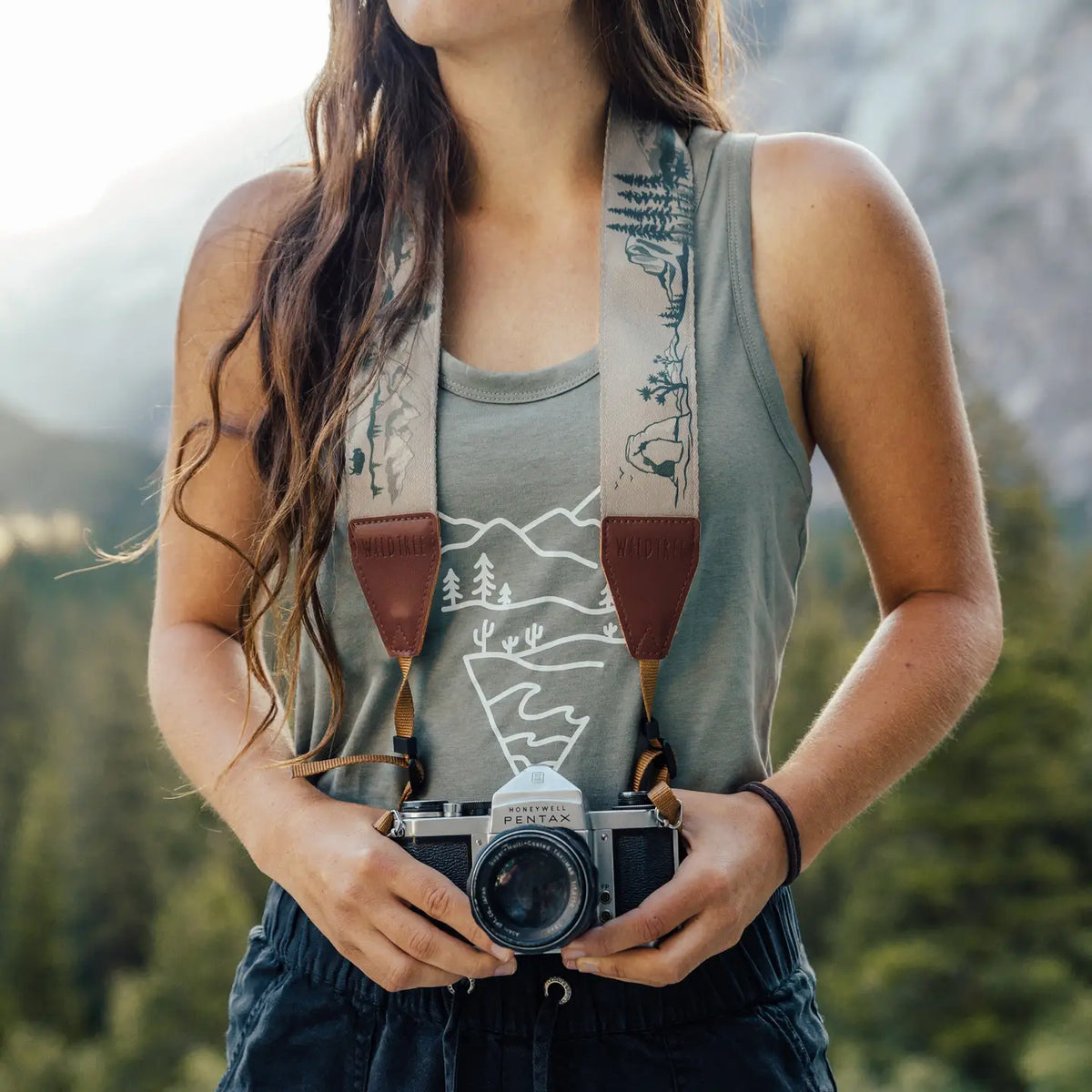 Outdoorsy person in tank top holding Wildtree National Parks Camera Strap, standing among green forest trees, adventure gear.