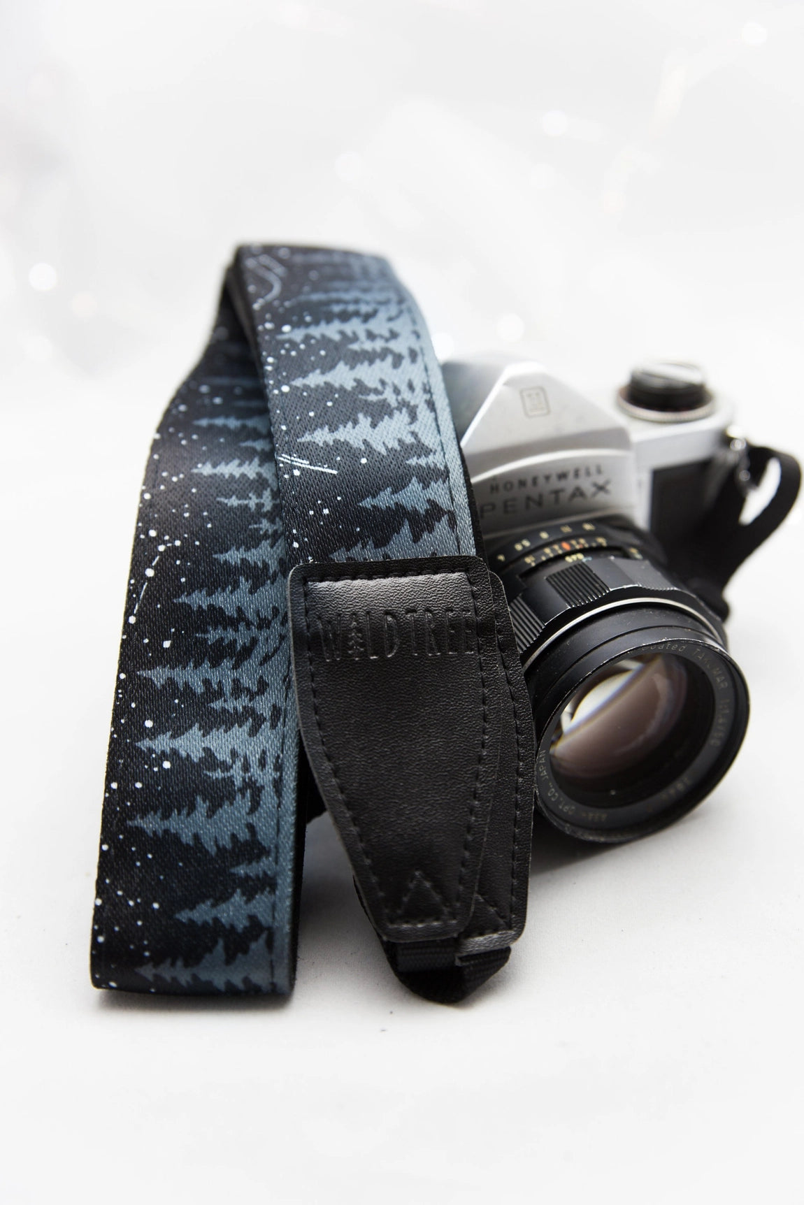 Film camera with celestial Wildtree Night Sky strap rests on white background; vintage photography gear, unique camera accessory.