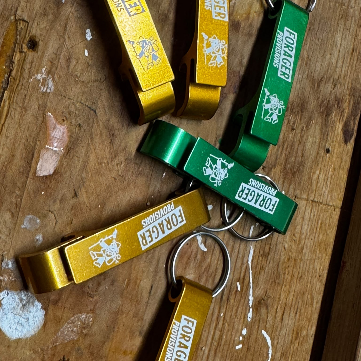Forager Provisions LTD bottle opener keyrings in orange and green with branded logos on a wooden surface.
