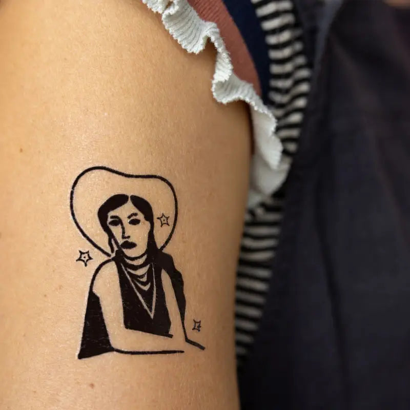 The Jenni Earle Cowgirl Dreamin' Temporary Tattoo, featuring a person wearing a wide-brimmed hat depicted in black ink with small star designs, exudes western vibes on the arm of someone wearing a striped shirt. This non-toxic tattoo is perfect for adding that touch of rustic charm.
