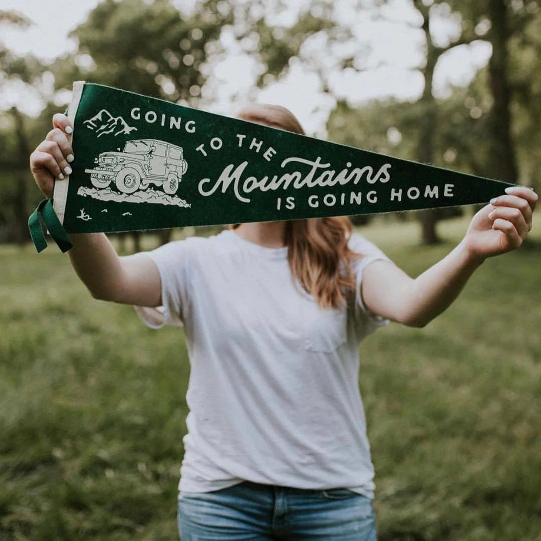 A person holding an "Oxford Pennant Going To The Mountains," which features the famous John Muir quote "Going to the Mountains is Going Home" along with a drawing of a mountain and vehicle.