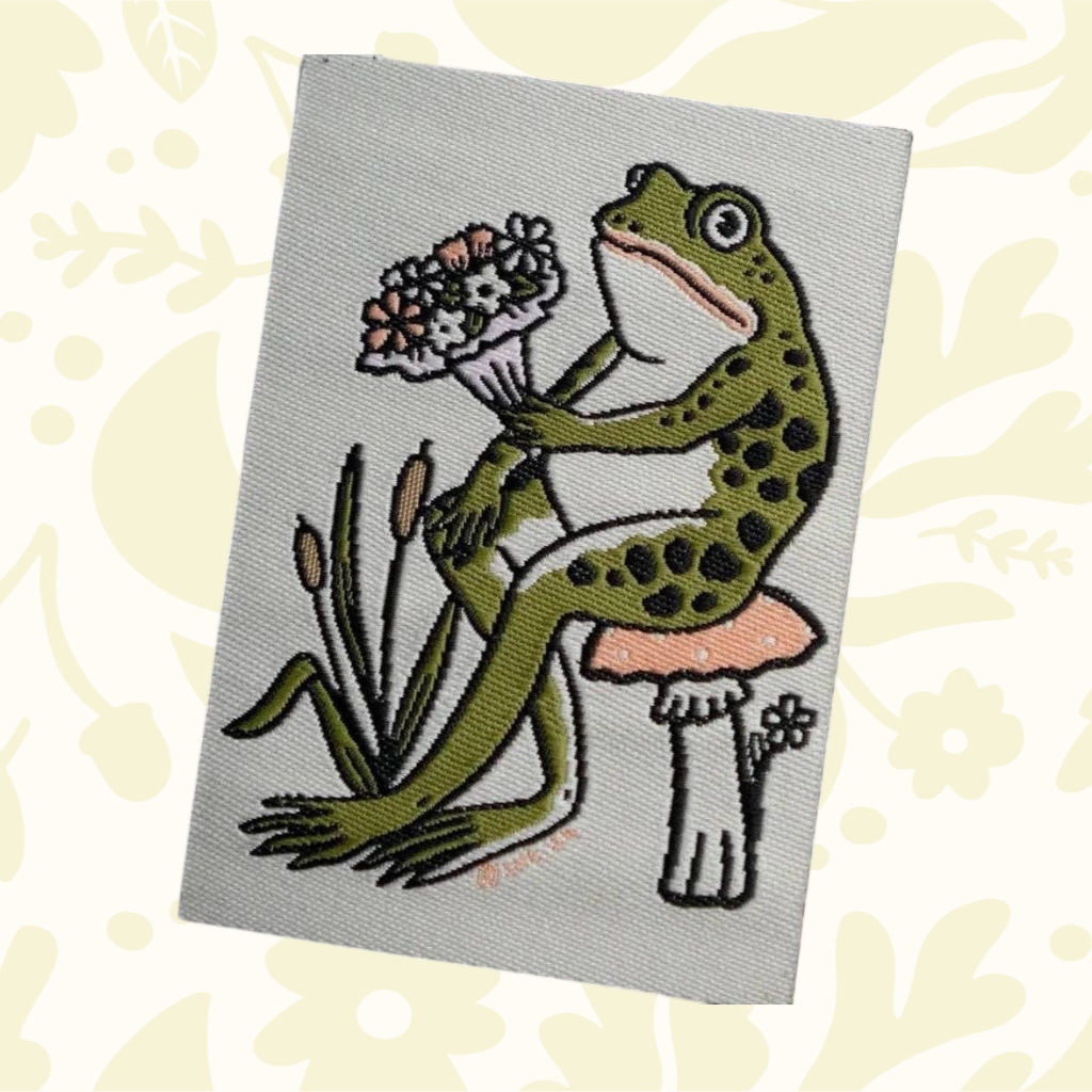 Forager Provisions LTD woven patch featuring a green frog sitting on a mushroom and holding a bouquet of flowers.