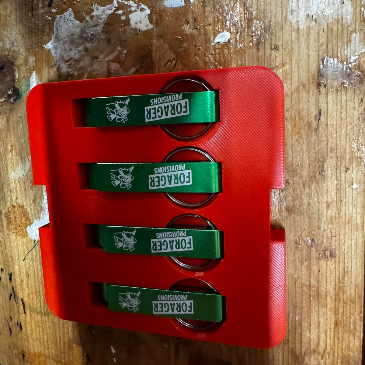 Forager Provisions LTD metallic green bottle opener keyrings in a red 3D-printed display tray on a wooden surface.
