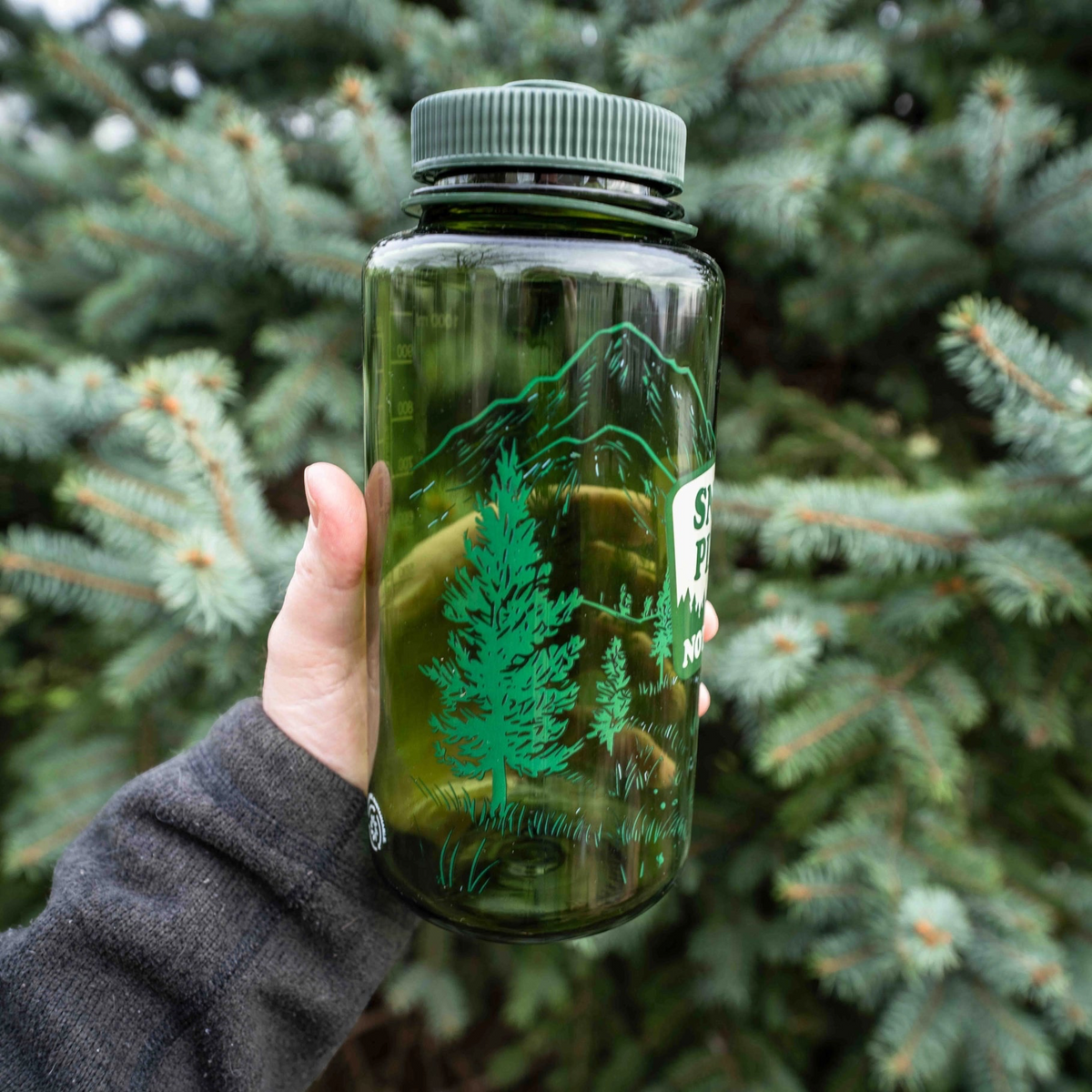 Forager Provisions LTD green Nalgene 32oz water bottle with Snort Pines Not Lines graphic held in front of pine trees