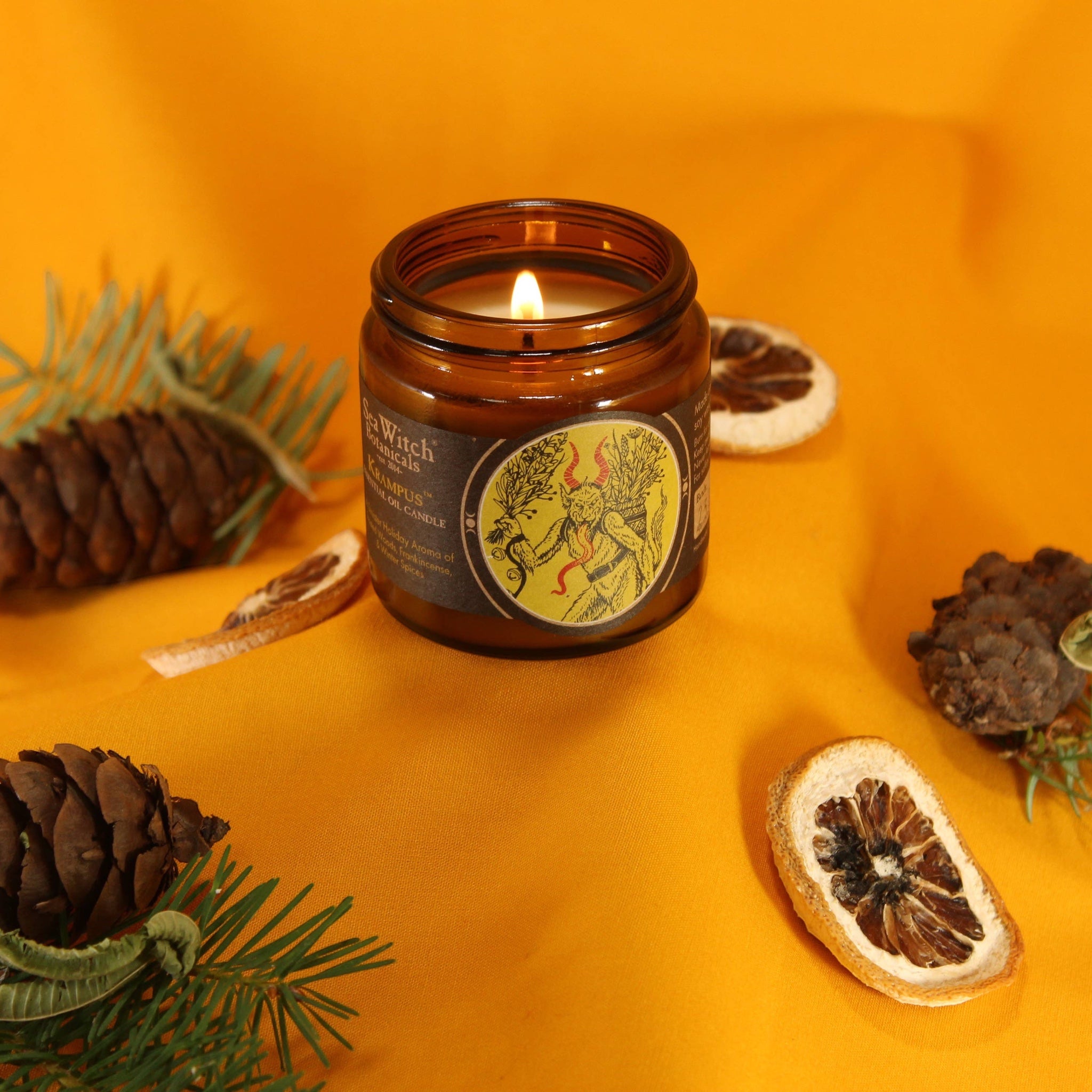 Forager Provisions LTD Sea Witch Botanicals Krampus soy wax candle in a glass jar, lit on a vibrant orange fabric.