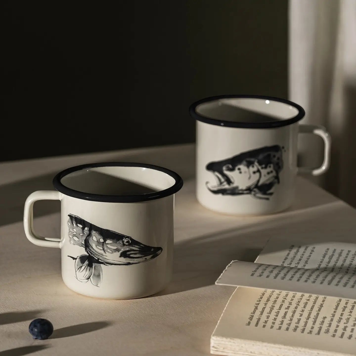 Muurla Teemu Järvi 0.37L Enamel Mug Pike & Trout on table with open book, fresh blueberry—unique Nordic homeware style.