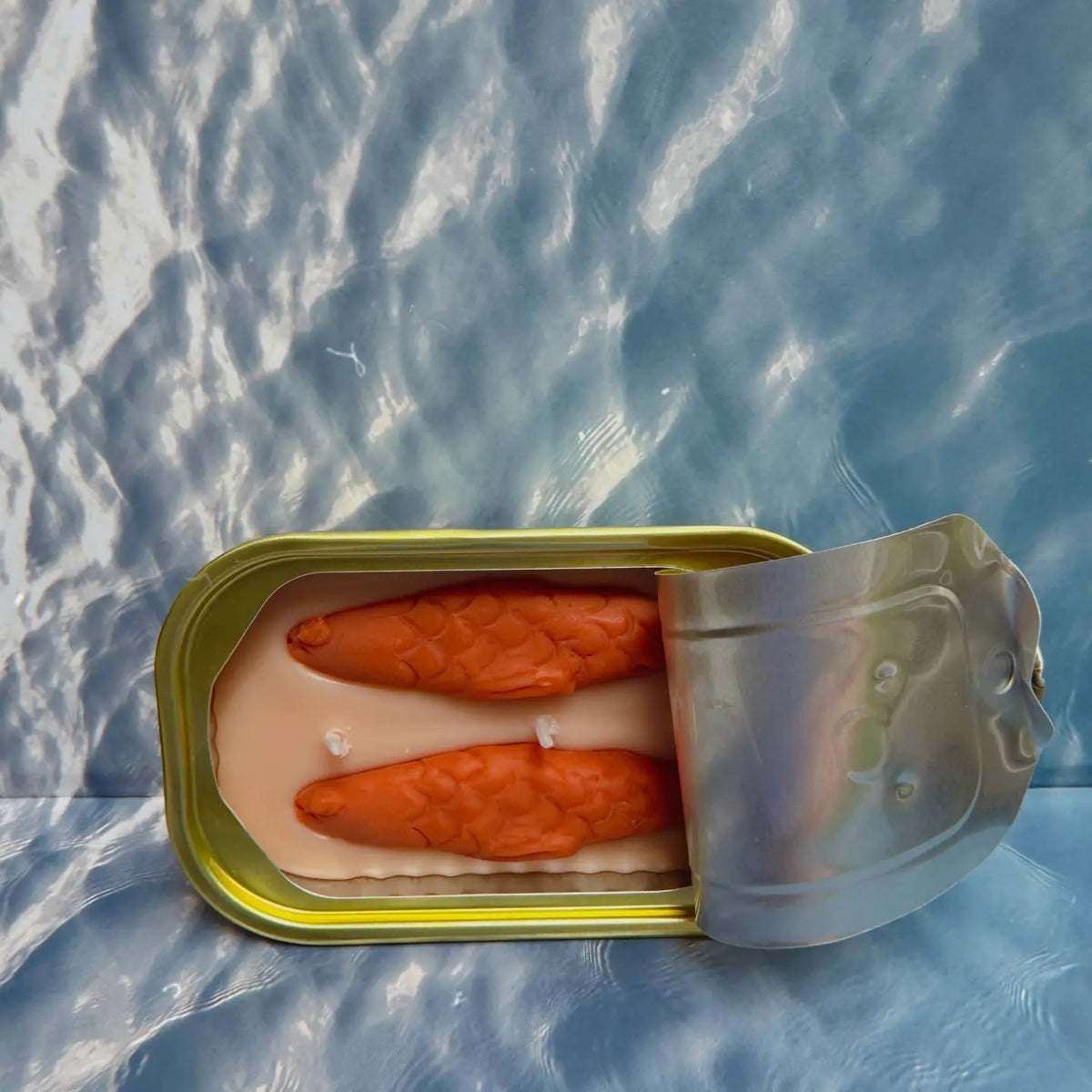 Forager Provisions LTD The Original Tinned Fish Candle with orange fish-shaped wax in a sardine-style tin on water background