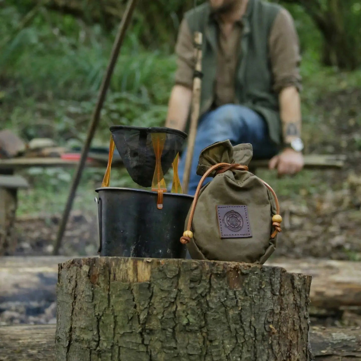 The Journeyman Craft Waxed Canvas Coffee Pouch in Khaki Green rests on a stump outdoors; blurred figure in the background.