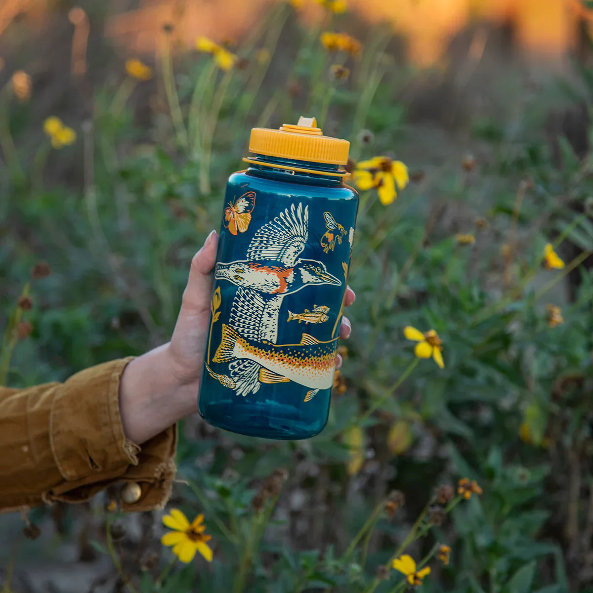 A person holds a blue water bottle with yellow lid, decorated with fish, plants, butterflies. Wildflowers and green foliage in background.