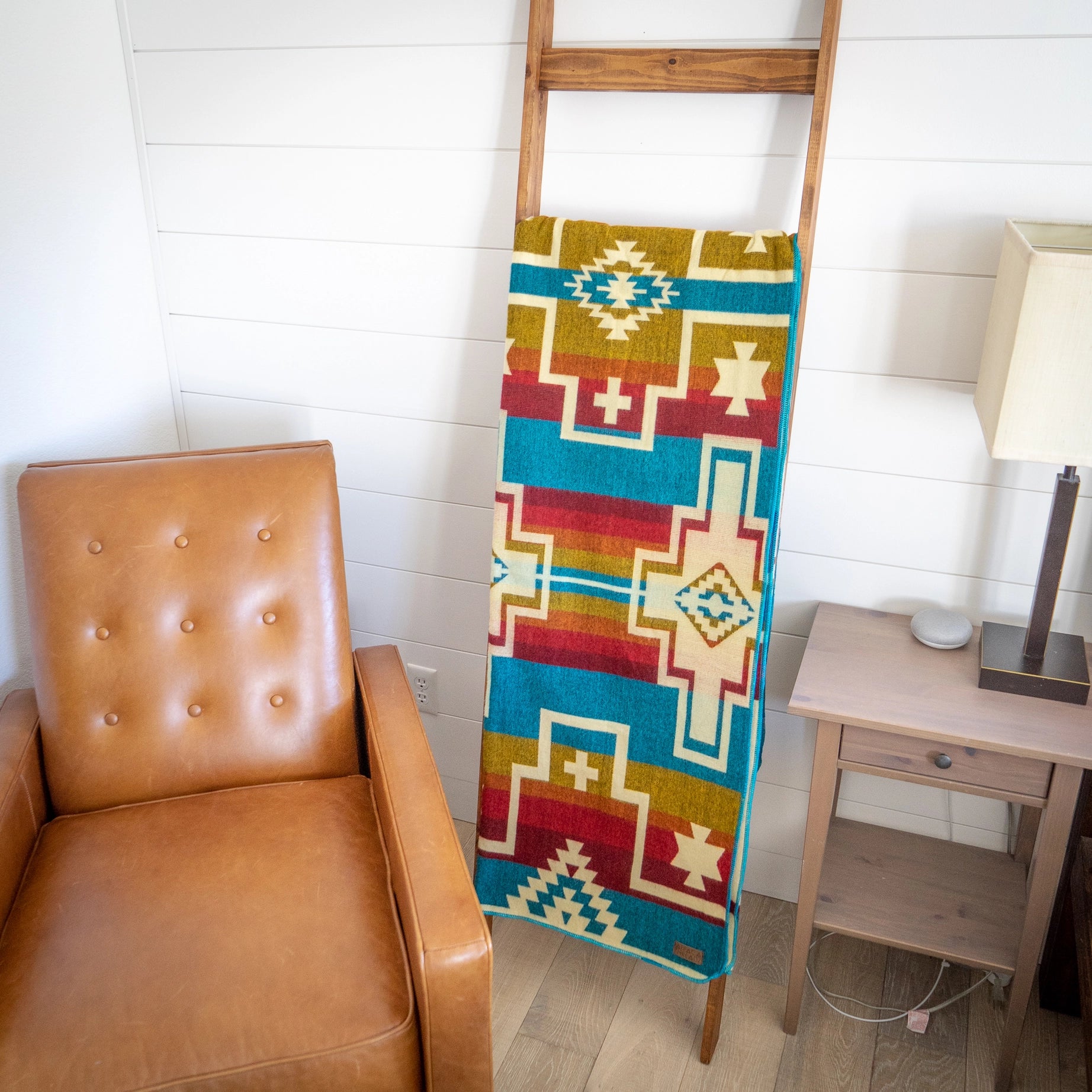 The Alpaca Threadz Wool Blanket Santa Fe, featuring a vibrant Native American pattern and handcrafted in Ecuador, is elegantly draped over a wooden ladder beside a leather chair and a wooden table with a lamp.