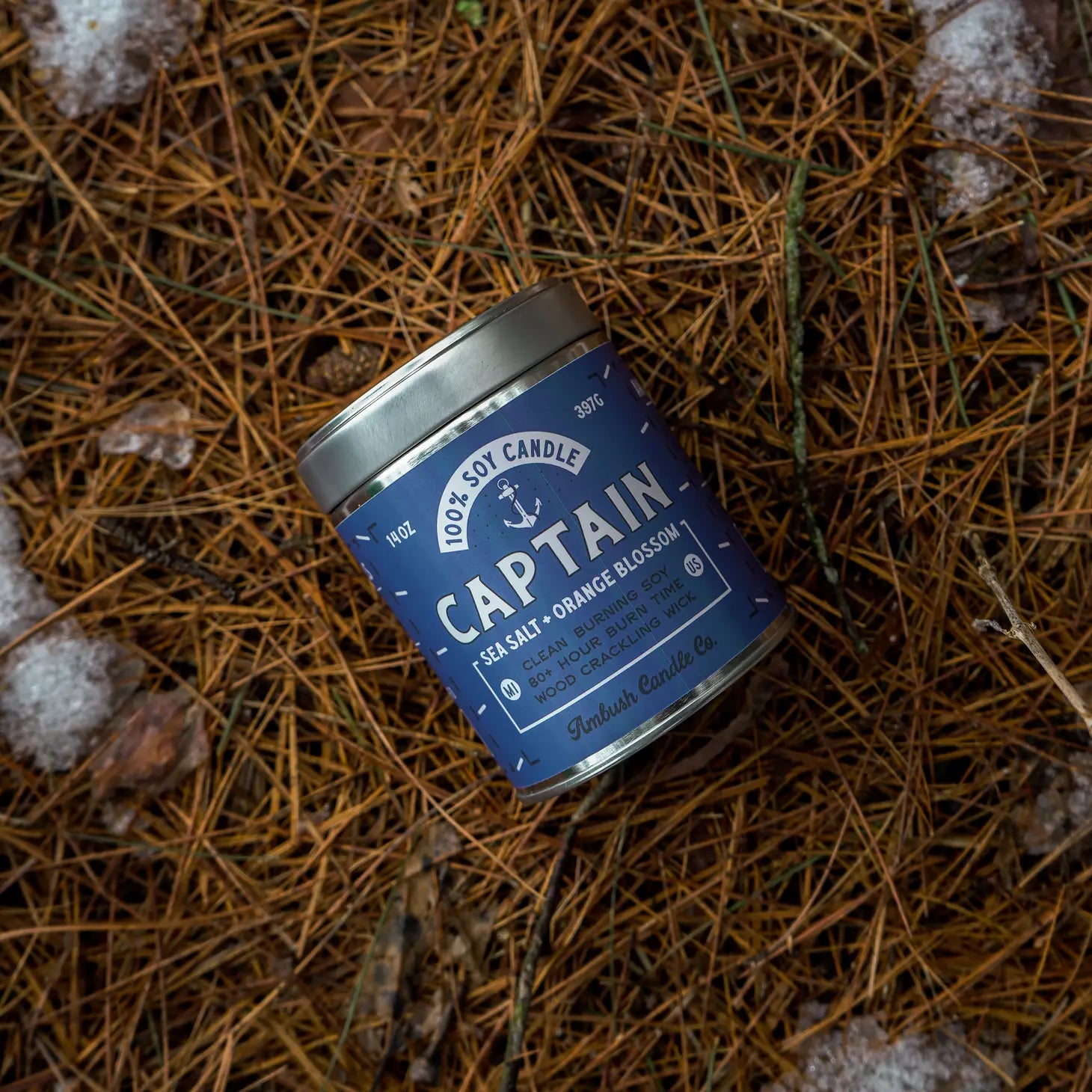 A tin of Ambush Candle Co Captain 14OZ Sea Salt & Orange Blossom Candle, placed on a bed of pine needles.