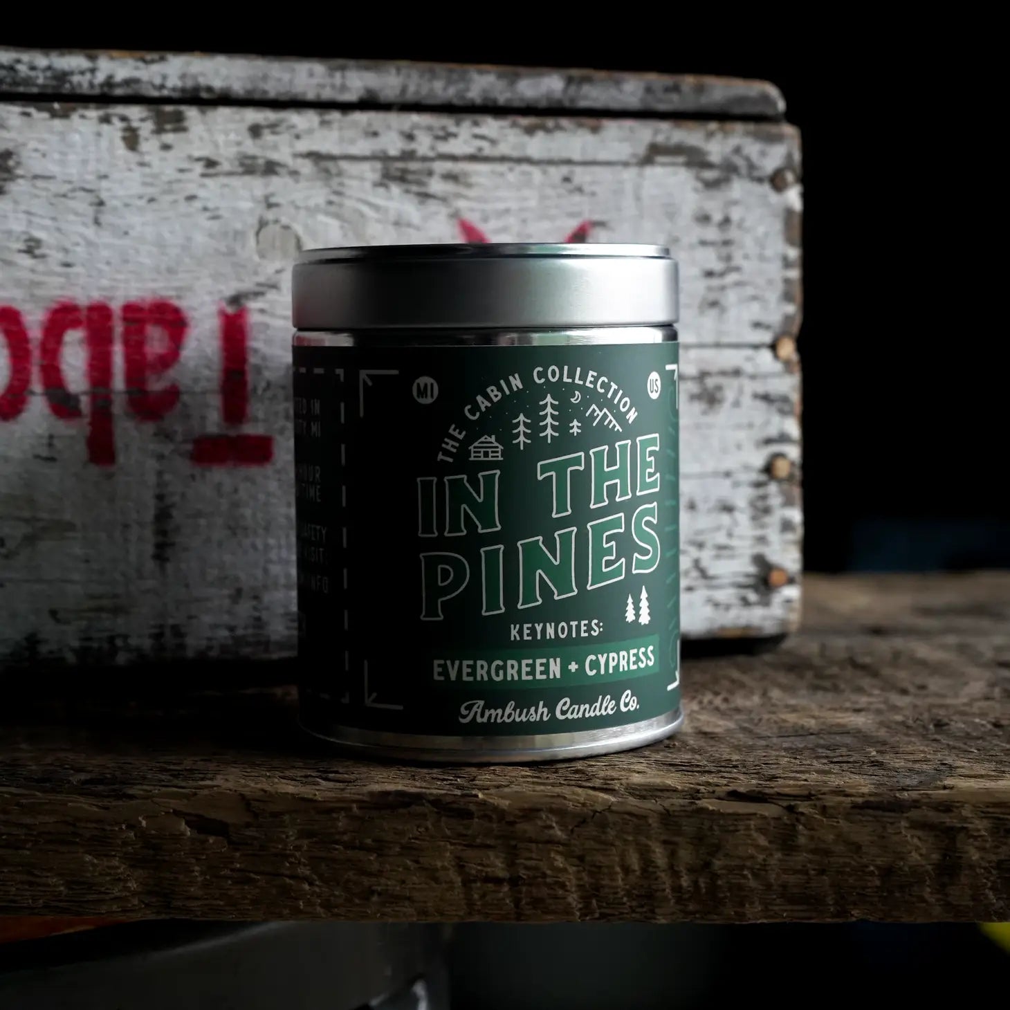A 14-ounce tin candle labeled "In The Pines Evergreen & Cypress" from Ambush Candle Co., made with soy wax, features notes of evergreen and cypress; the lid is off to the side.