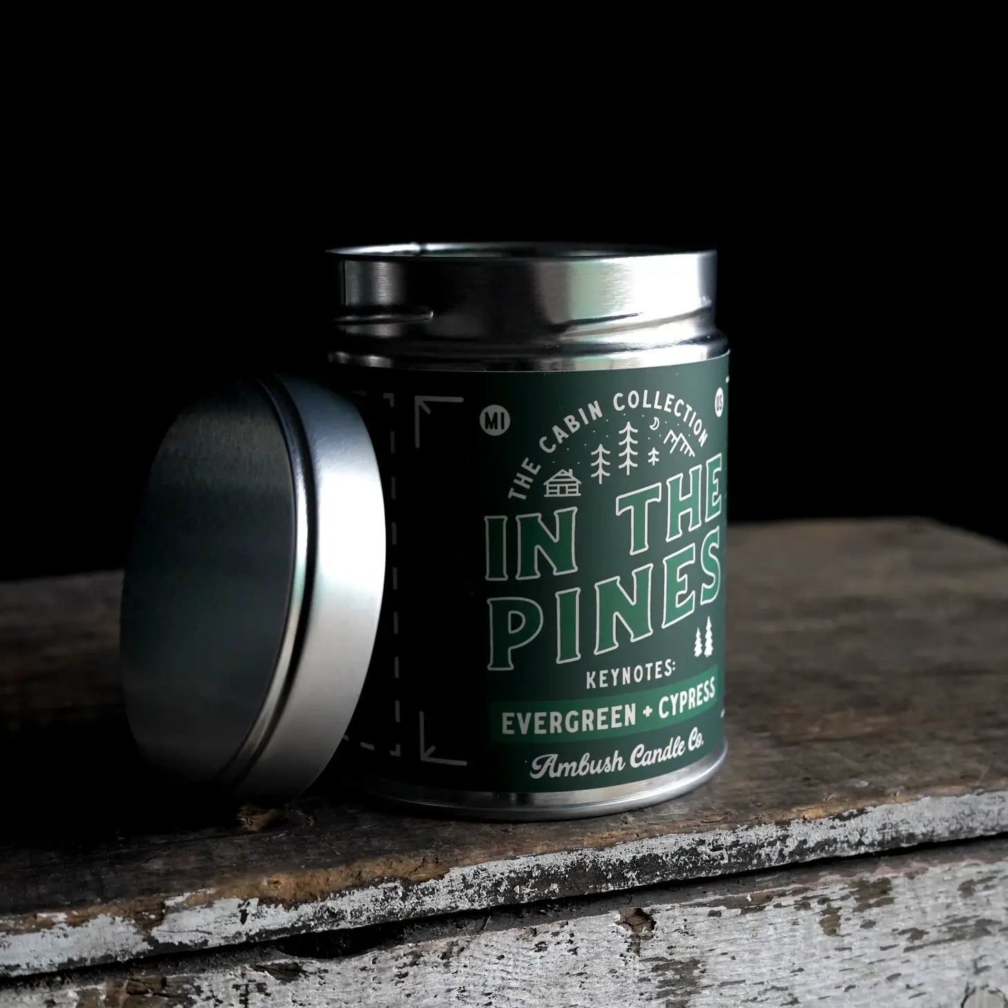 A 14-ounce tin candle labeled "In The Pines Evergreen & Cypress" from Ambush Candle Co., made with soy wax, features notes of evergreen and cypress; the lid is off to the side.