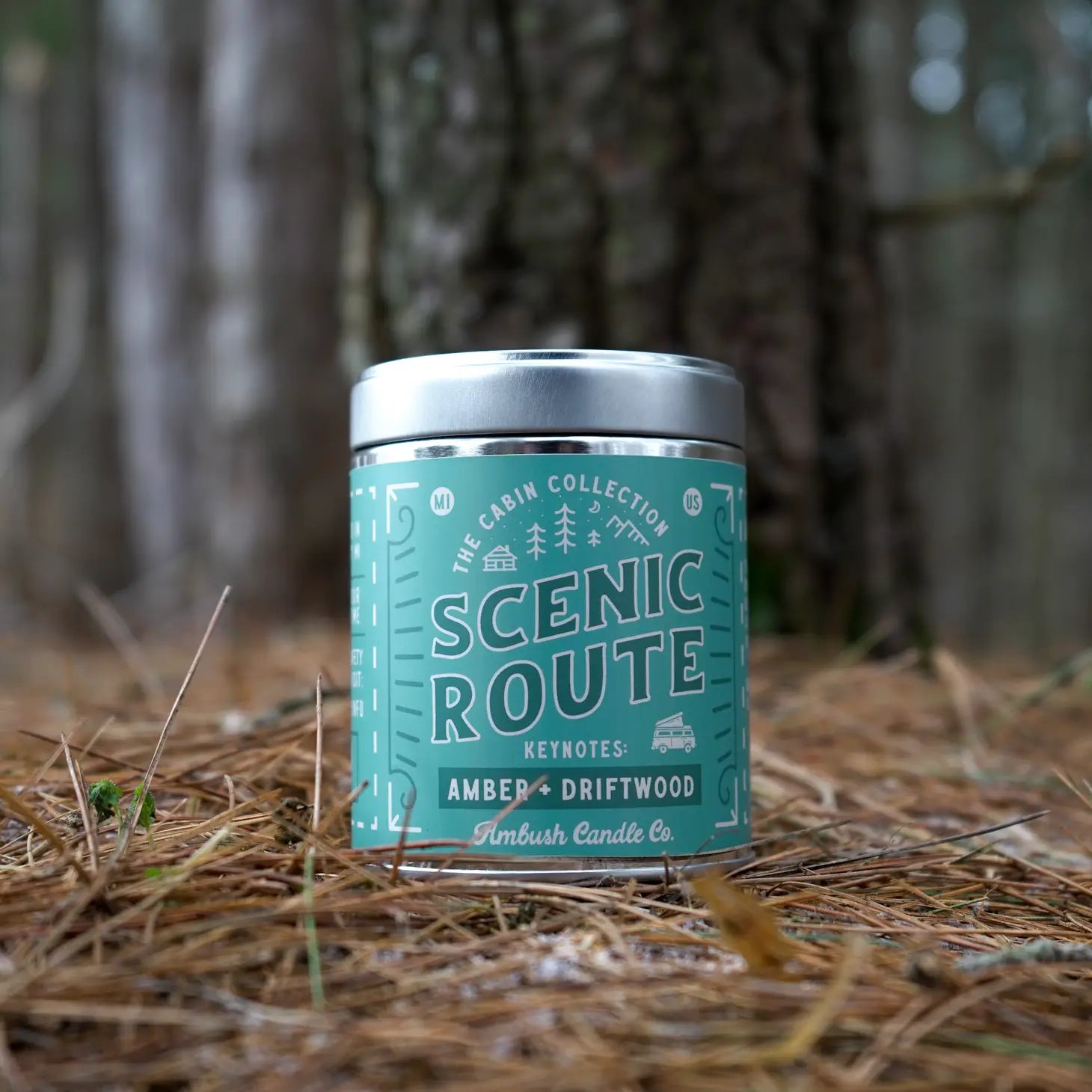 Three candles are stacked outdoors, with the top candle labeled "Ambush Candle Co 14OZ Scenic Route Amber & Driftwood Candle," made from eco-friendly soy wax.