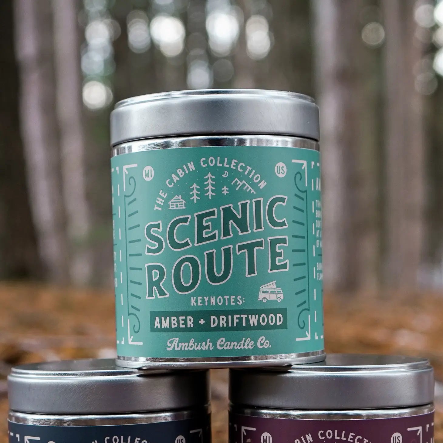Three candles are stacked outdoors, with the top candle labeled "Ambush Candle Co 14OZ Scenic Route Amber & Driftwood Candle," made from eco-friendly soy wax.